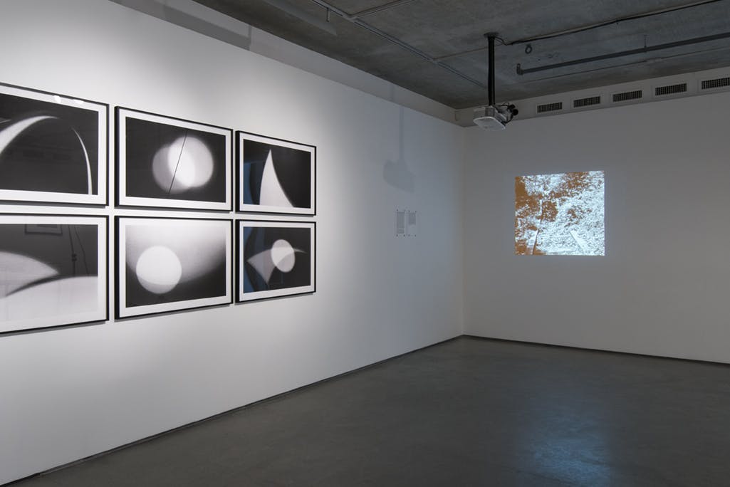 A series of black and white abstract photographs are installed on a wall. A single-channel video of a landscape is projected on the other wall.