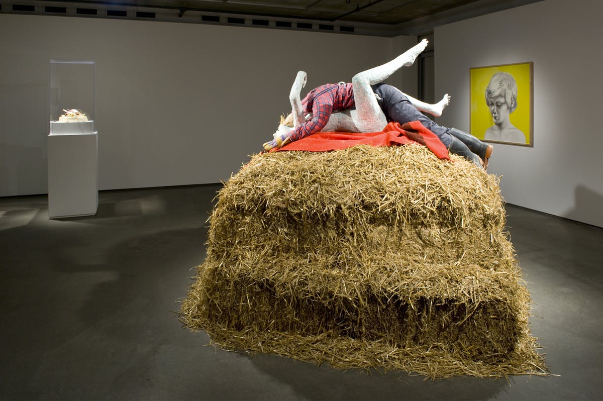 A large pile of straw sits on a gallery floor on which two, embracing mannequin-like forms are laid. A small sculpture under glass on a plinth and a portrait-style framed artwork are visible behind.