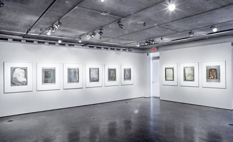 In a gallery with concrete floors and ceilings, there are nine framed photos. The photos each show one charred and damaged book. The bokos are all different colours, shown with a grey background.