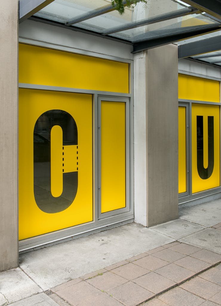 Detail image of Kay Rosen’s work on CAG’s exterior facade. Yellow vinyl covers the windows. A capital letter “C” has been cut out of the vinyl on one window, with a “U” cut from the window next to it.