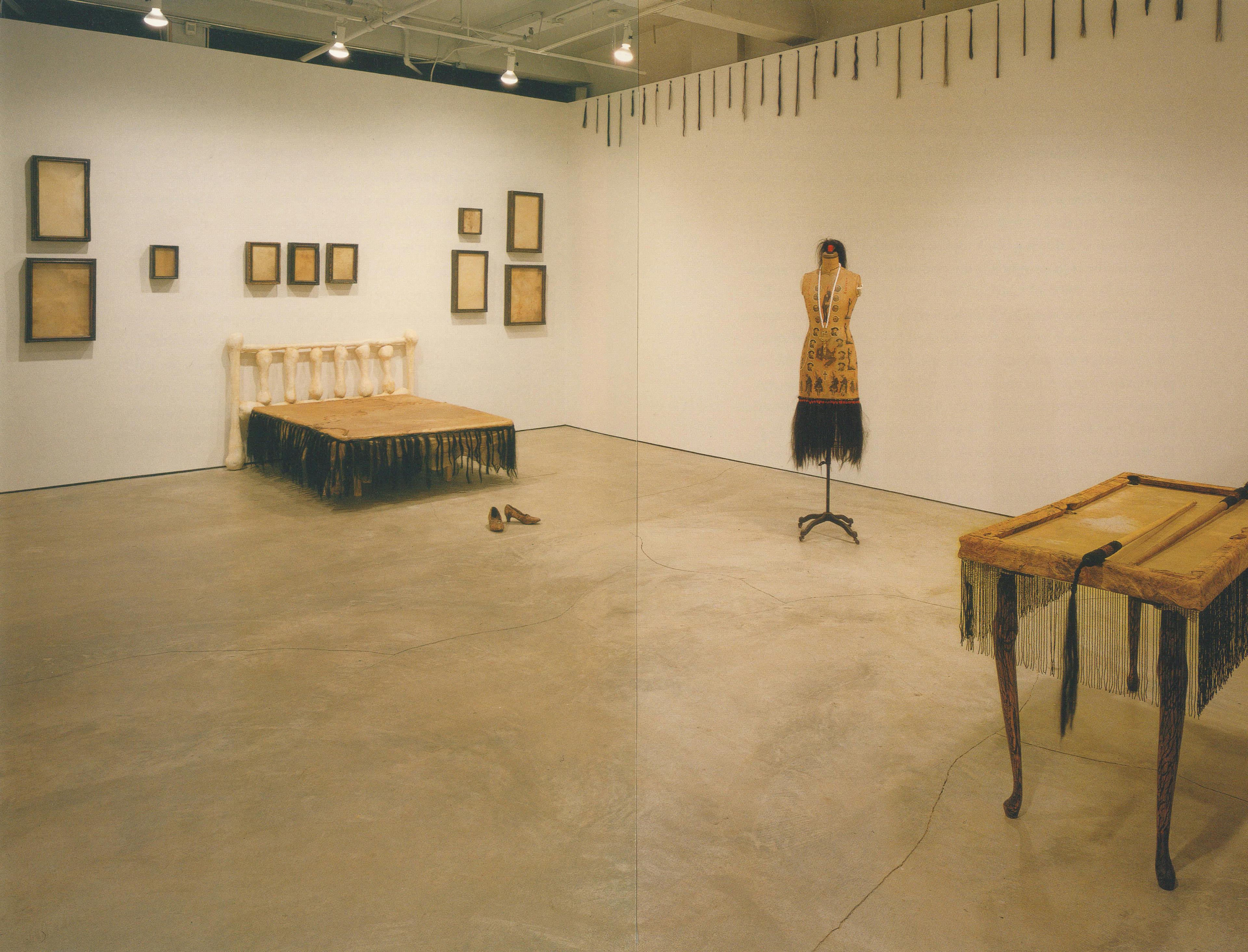 Many brown-coloured artworks are installed in a gallery. A wood table, a dressmaking mannequin, a pair of shoes, and a bed are placed on the floor. Ten paintings are mounted on the wall above the bed.