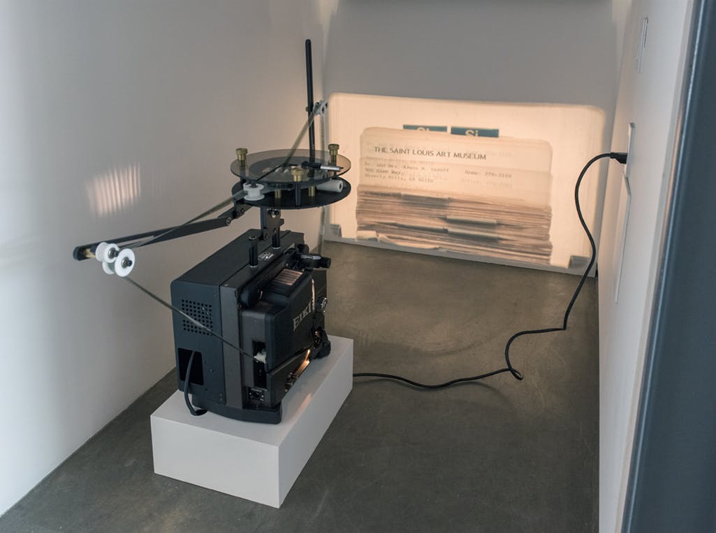 A black projector is installed in a low alcove, projecting an image on the white wall. The image depicts a rolodex of cards. One of the cards reads: THE SAINT LOUIS ART MUSEUM. 