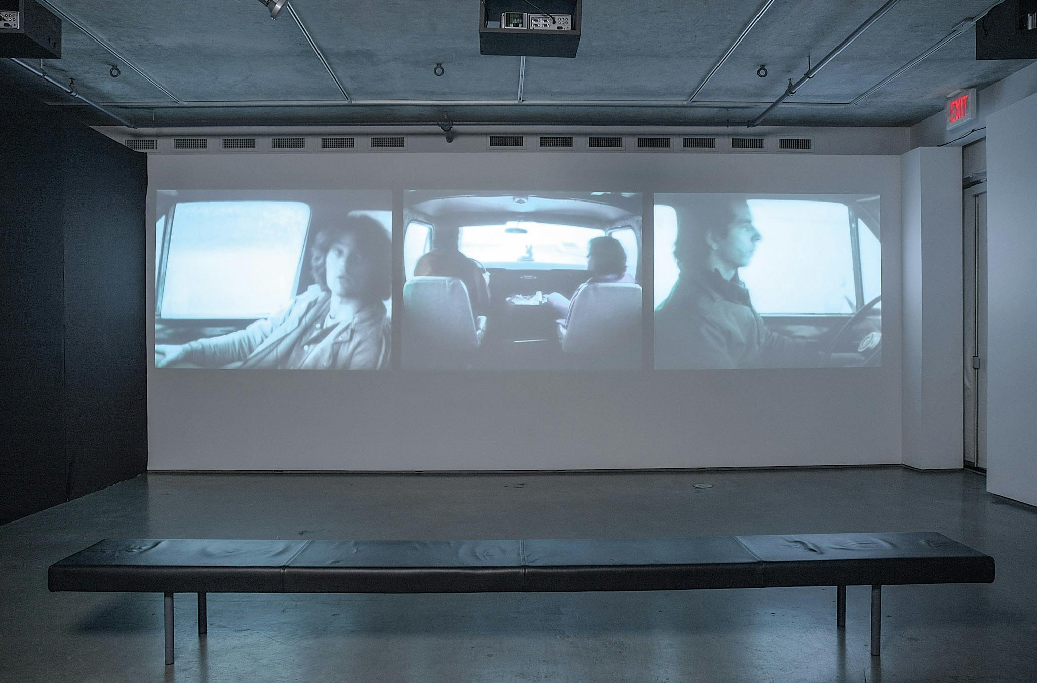 An installation image of three-channel video work projected on a gallery wall. The videos show people in a car from different angles. The middle video shows an image of two people in front seats.