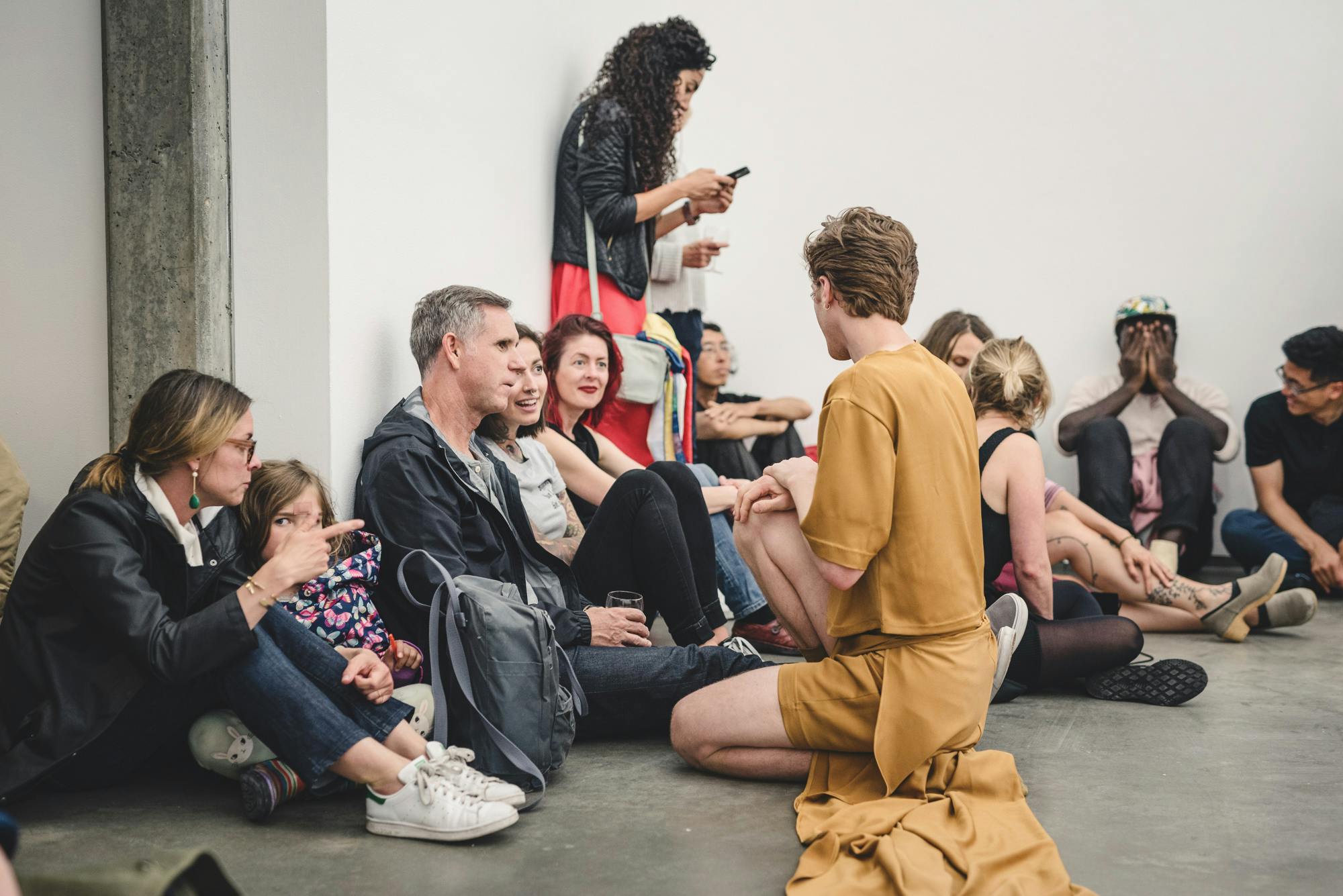 Andrew Bartee, wearing a mustard-coloured outfit is kneeling down on the gallery floor to talk to some of the audience sitting besides the walls. 