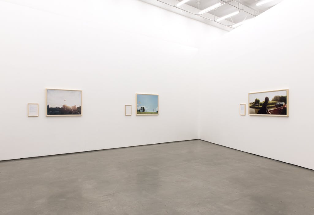 Three framed, colour photographs are installed on adjacent gallery walls. Each is an outdoor scene. A framed text is installed to the left of each photograph.