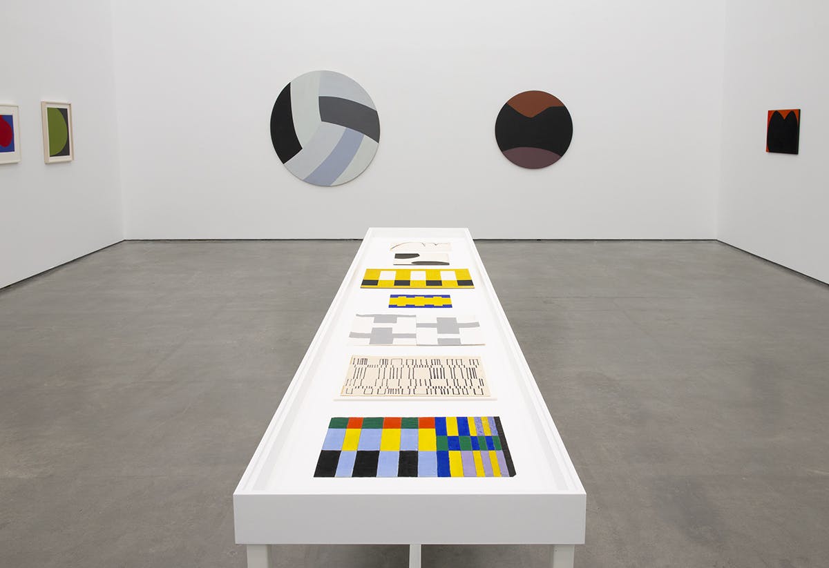 A vitrine sits at the center of a gallery space surrounded by walls displaying the paintings of Leon Polk Smith. A series of small colourful geometric paintings lay flat in the vitrine.