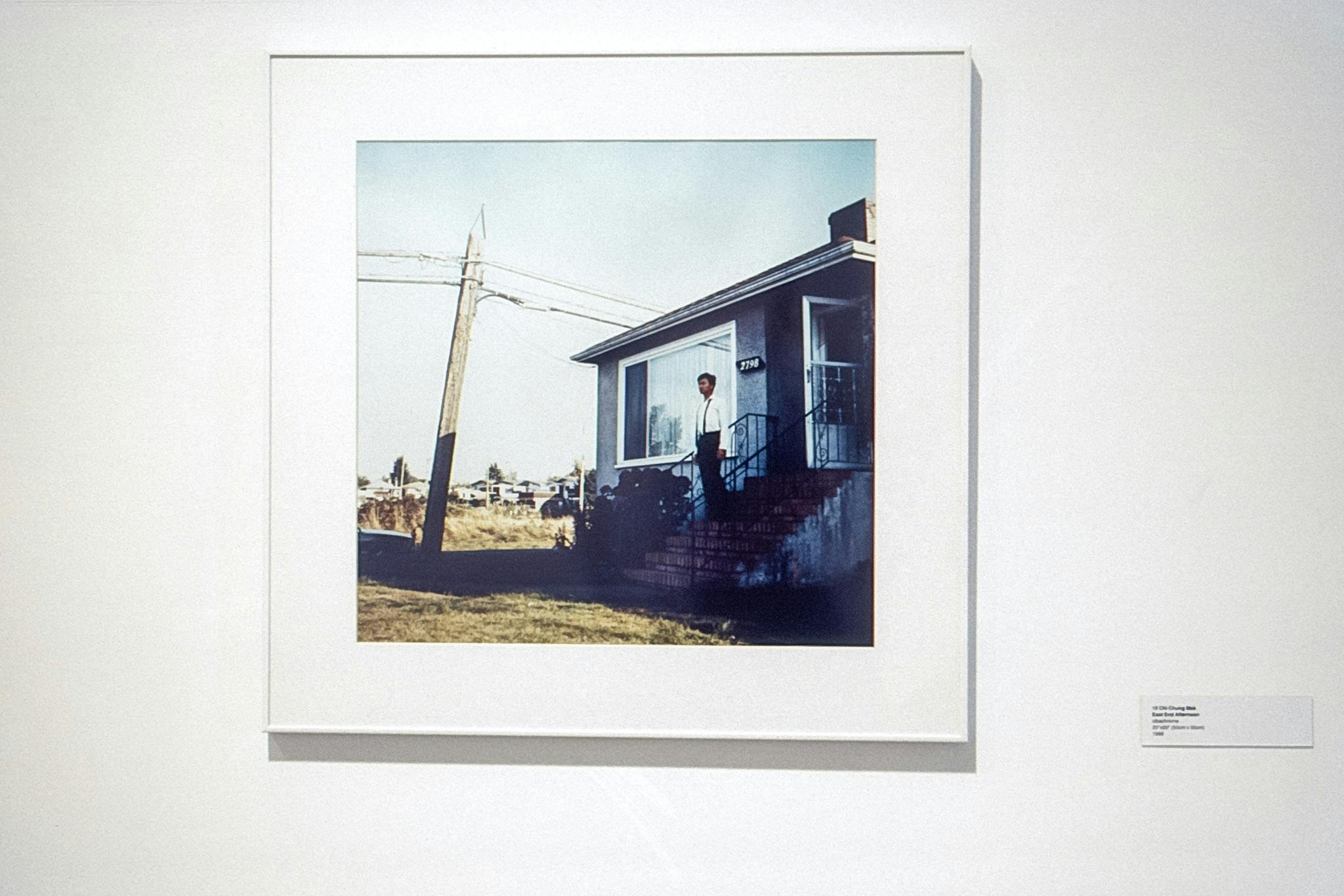 A closeup of a colour photo in a white frame, on a white wall. The photo shows a person standing on the steps of a grey, single-story house, with a small telephone pole just on the side of their lawn.