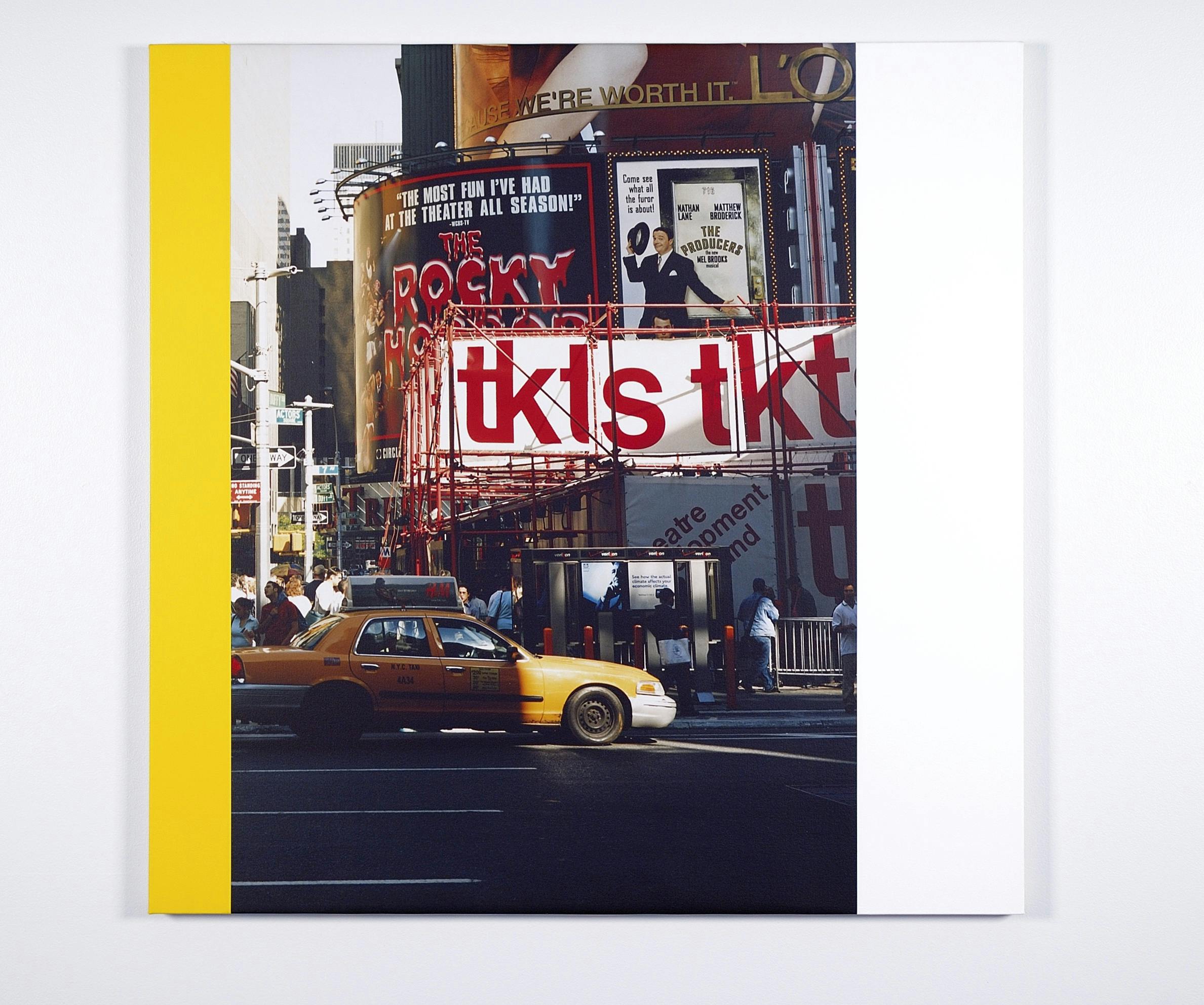 A photograph of a busy city street corner hangs on a white gallery wall. A strip of yellow paint runs down the left side of the photograph. 