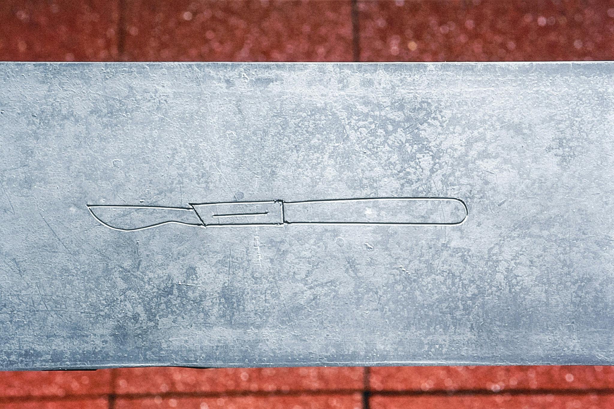 A shape of a small carving knife is carved on the metal surface above the floor covered by the rubber tiles. The depicted knife looks skinny and half of its entire body is made up of the handle. 