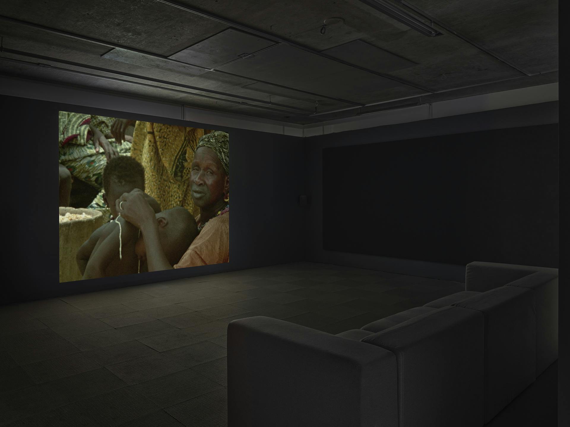 A video on a wall in a dark room depicting a person looking at the camera; a couch is visible in the foreground.