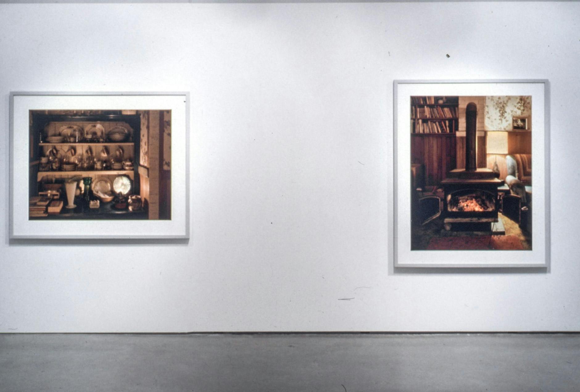 Two large-sized coloured photographs are installed on the gallery walls. Both of them depict the interior of a wood cabin. The one on the right shows a small fireplace beside bookshelves. 