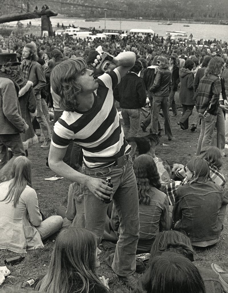 A black and white photograph depicting a person wearing a striped shirt drinking from a glass bottle. They are outdoors in a vast crowd of people milling around or seated in the grass.