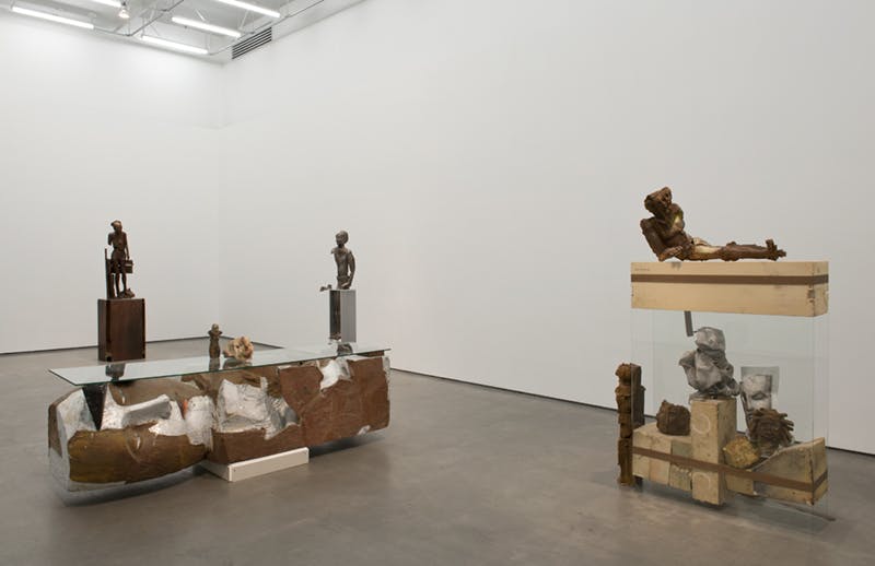 Variously sized sculptures installed on plinths inside a gallery. In total, twelve human form sculptures of different materials are visible. Some are copper coloured, and some are light grey.