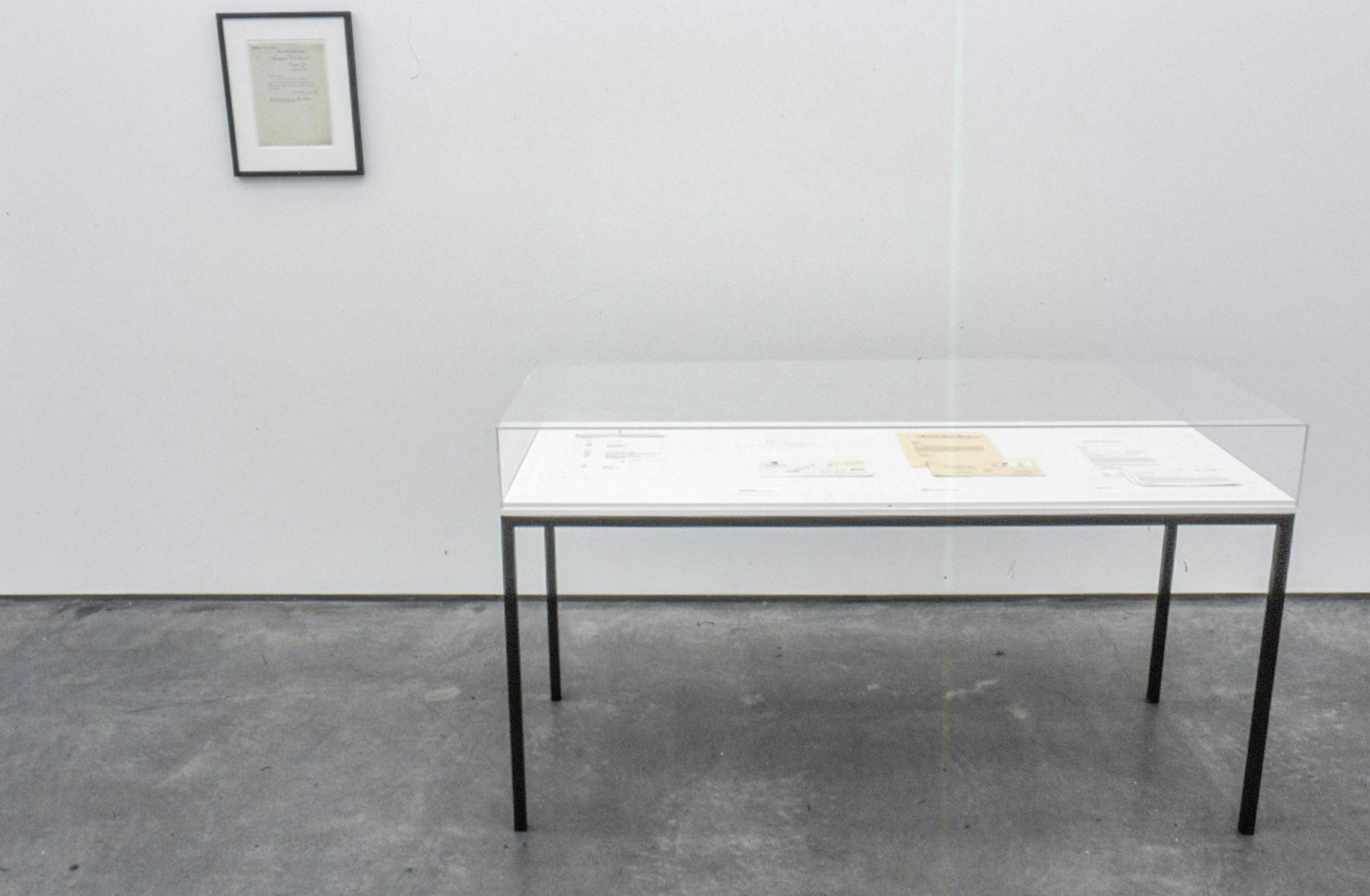 An installation image of Kirsten Pieroth’s art exhibition. Various pieces of paper with texts printed on them are placed in a display case. Another document is framed and mounted on the wall above the display case. 