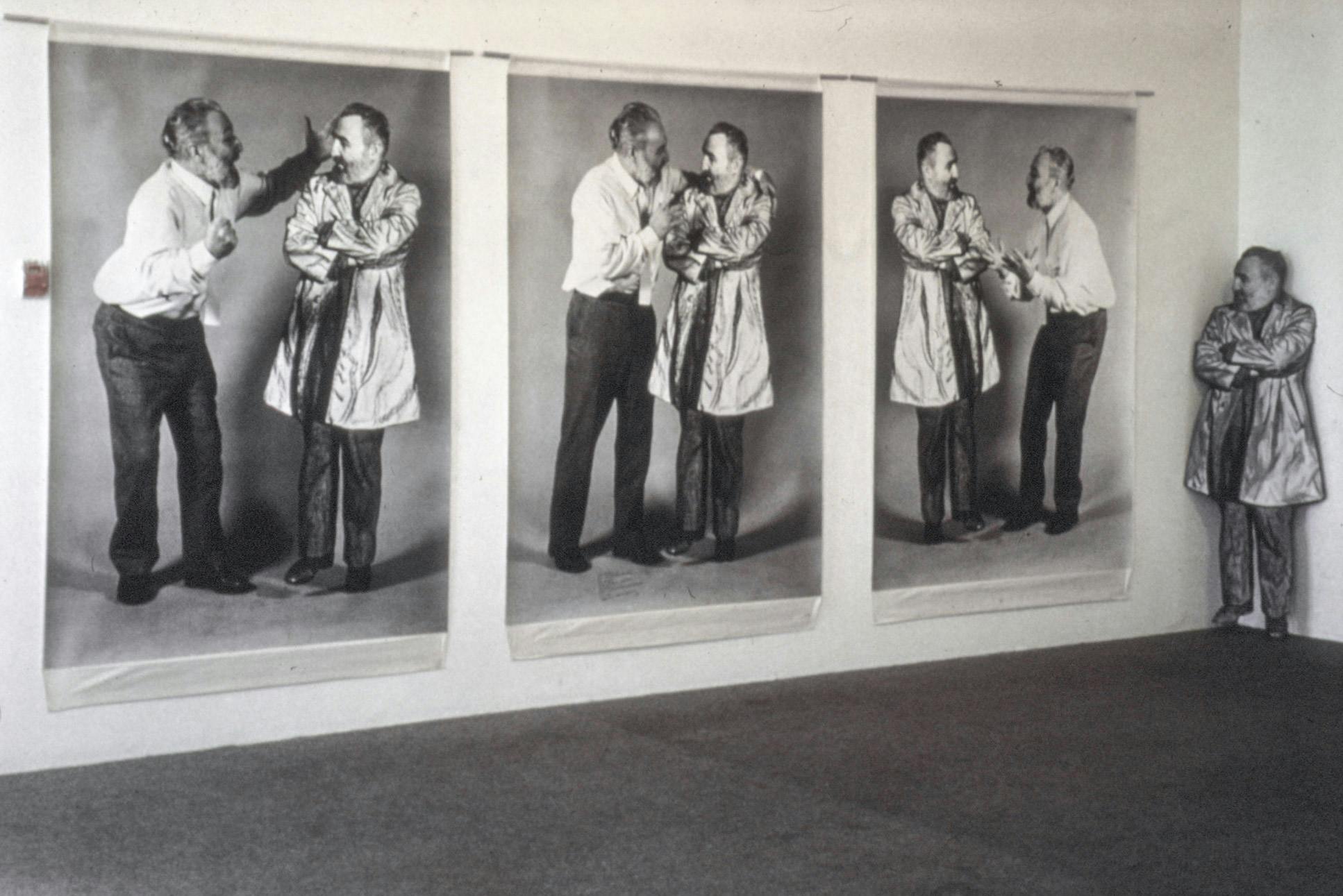 On a wall, there are 3 life-sized black and white photos of a person in business attire talking to a cardboard cutout of themself. The cardboard cutout is placed in the corner beside the photos. 