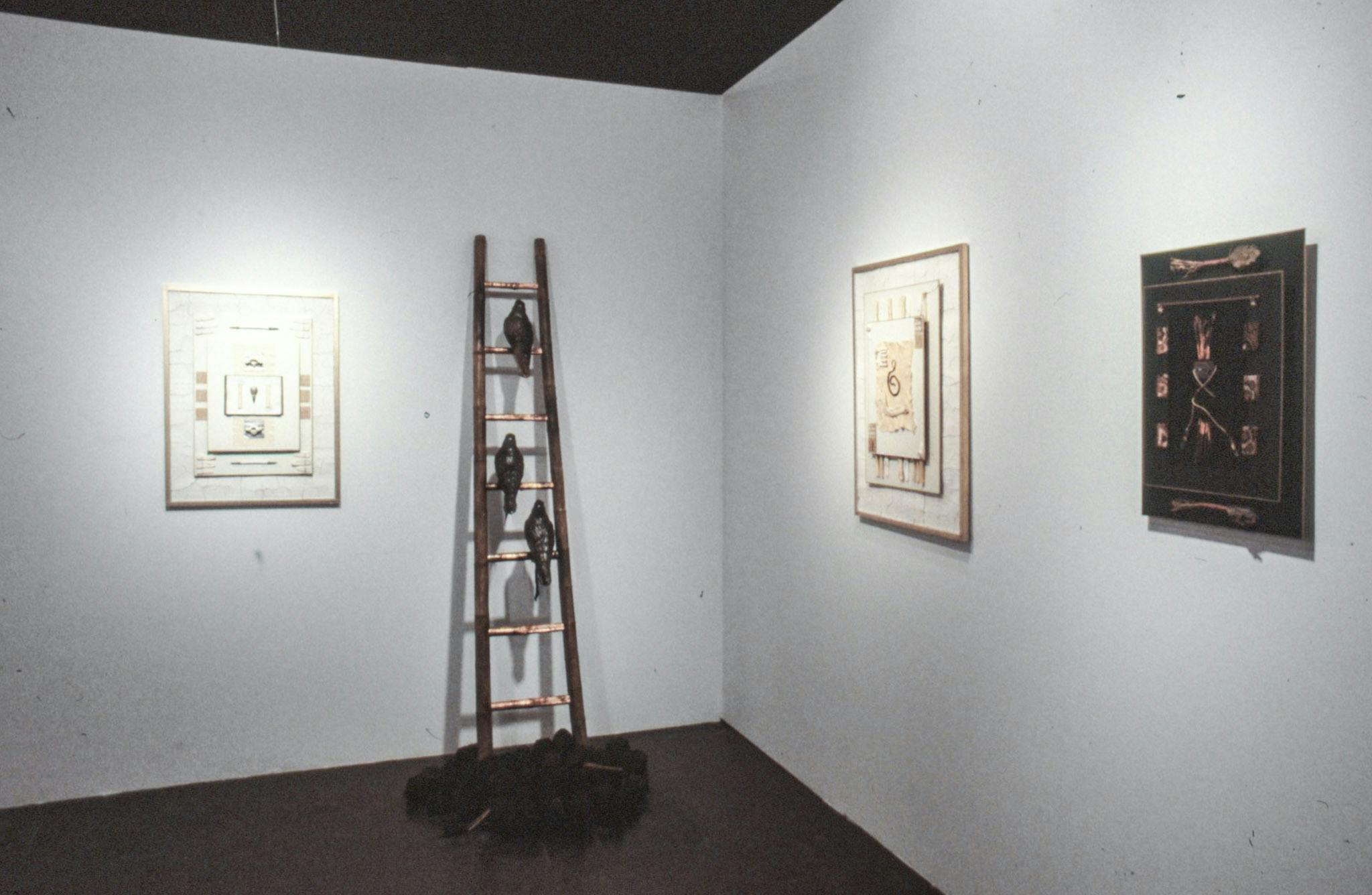 The corner of a gallery with several artworks on the wall. The works include framed collages, drawings, and leather pieces. One of the works is also a sculpture of a bronze ladder with birds.