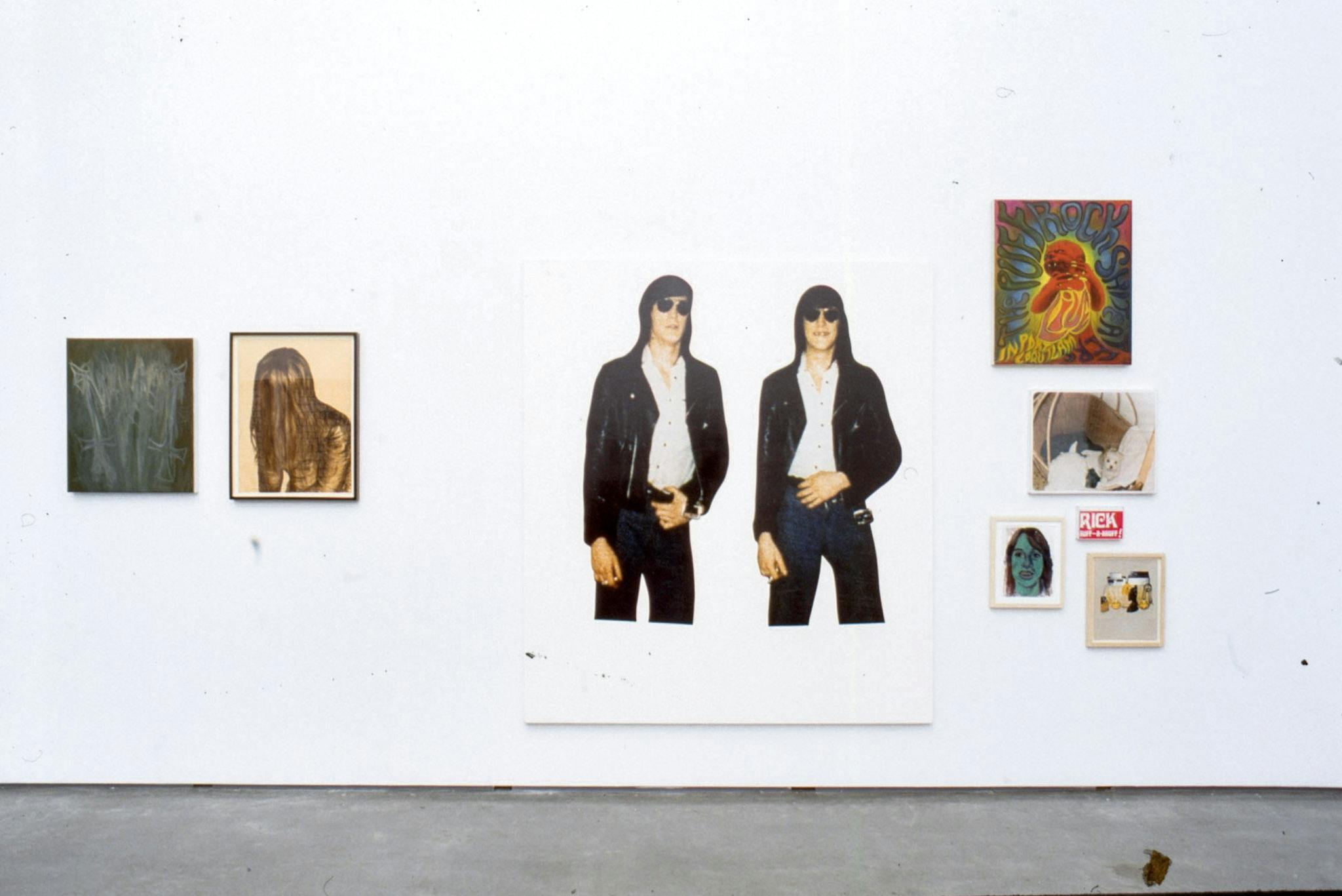 Installation image of works by Steven Shearer. Paintings, collage works, and photographs are mounted on the wall. Many of them are coloured. Some works show figures of a person with long straight hair. 