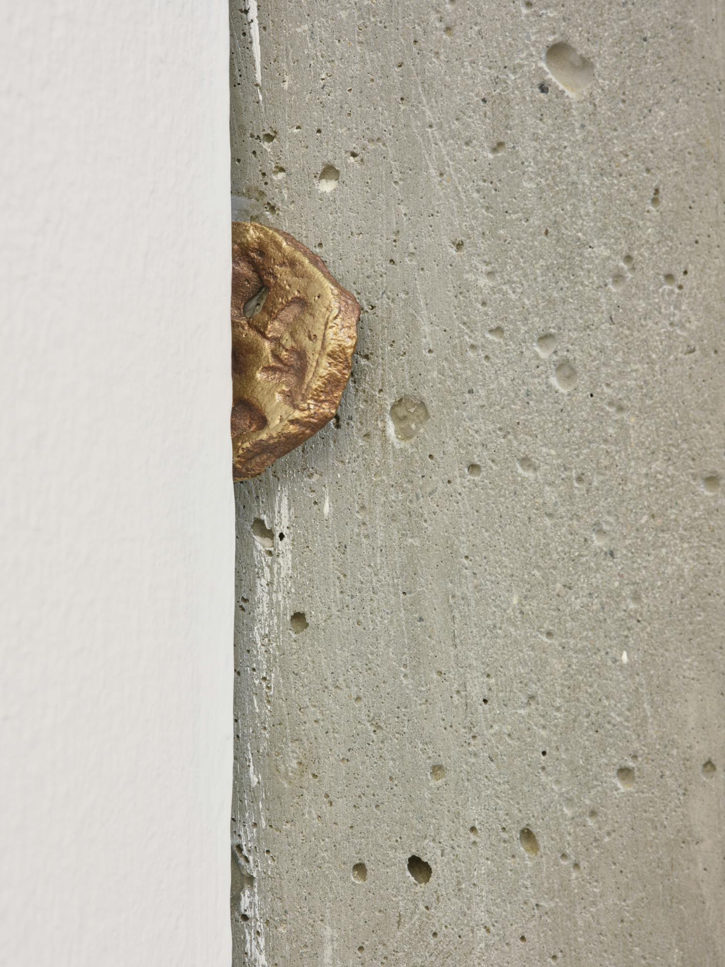 A small gold sculpture resembling a lotus root is wedged between a cement wall and white drywall.