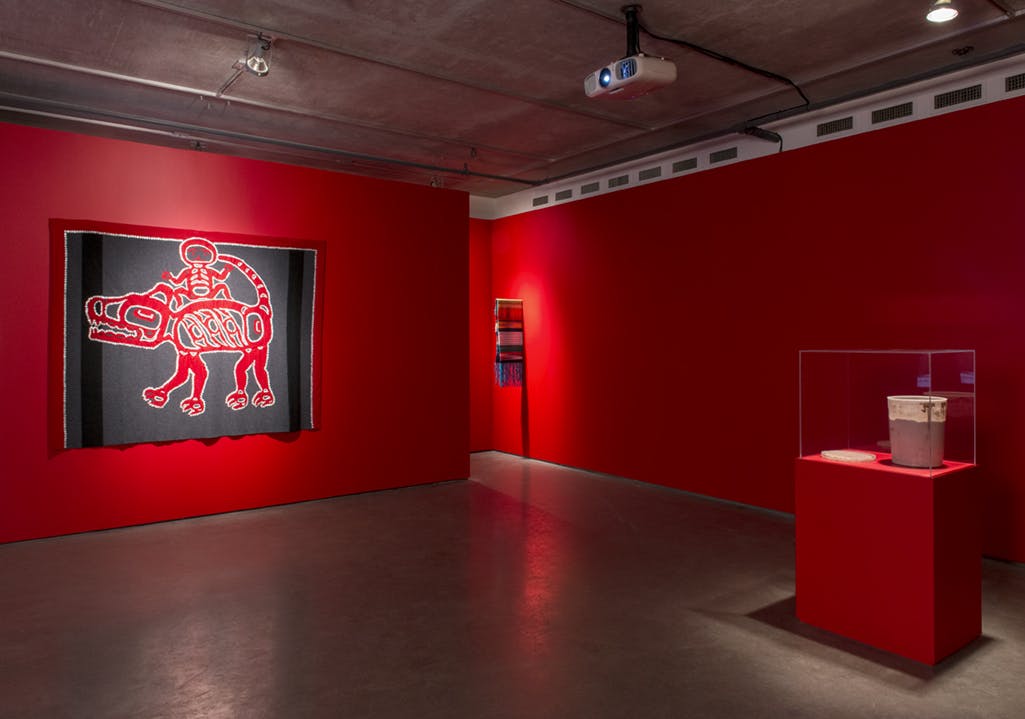 A large grey tapestry is installed on a red-painted gallery wall. The tapestry depicts a small human figure riding on a four-legged animal. 