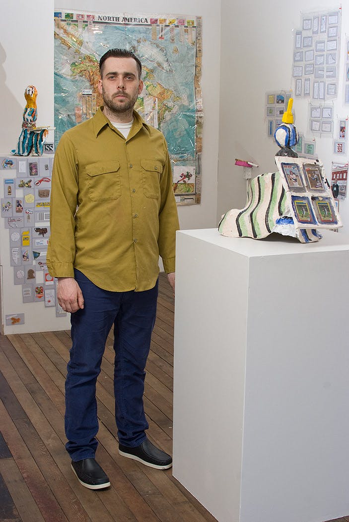A portrait of Mark DeLong. He is standing in a gallery space next to a plinth which displays a colourful mixed media sculpture.