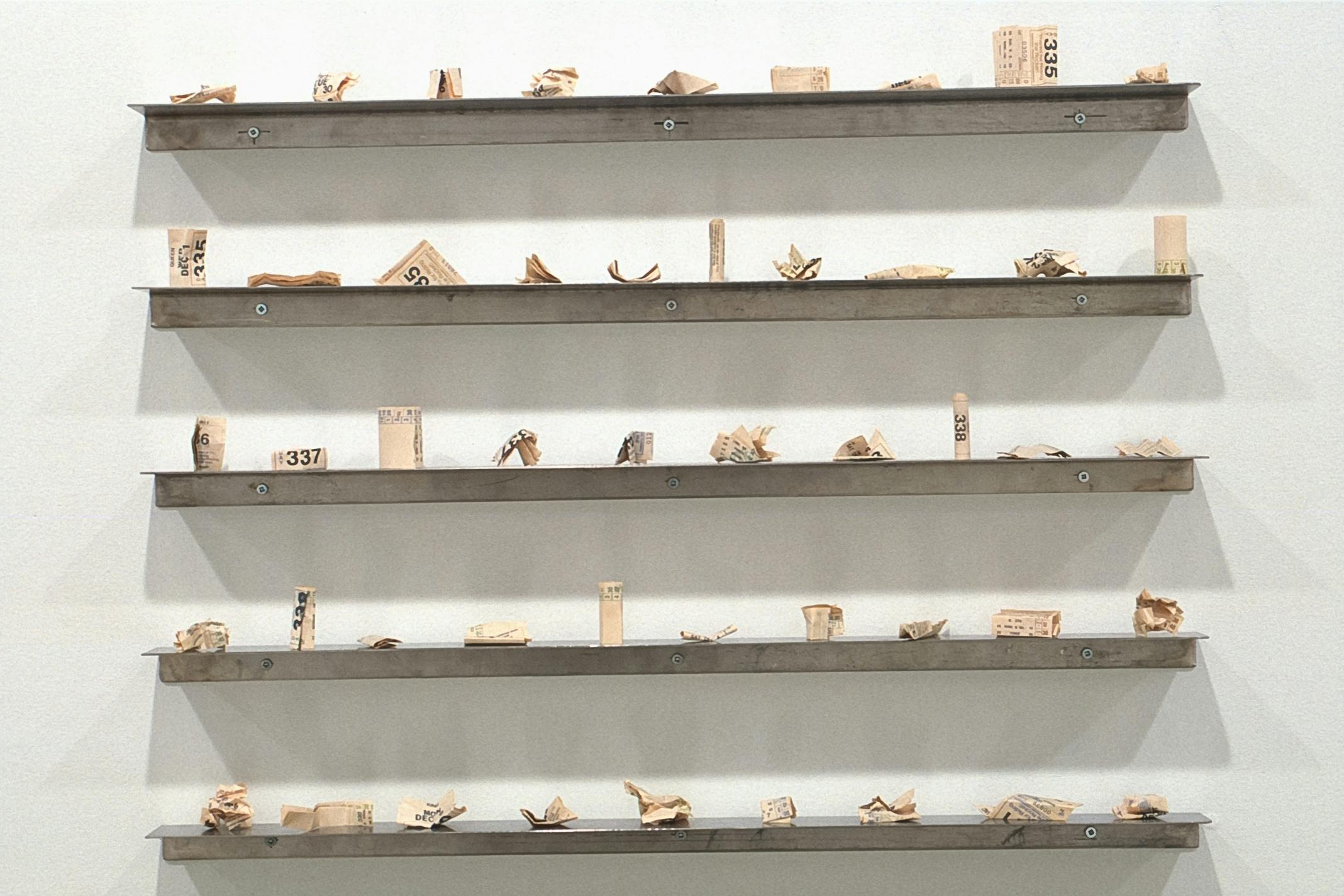 Small-sized sculptures are installed on the white gallery wall. They are all made out of crumpled paper. Some look more cylindrical than others. There are five rows of ten sculptures on the wall.