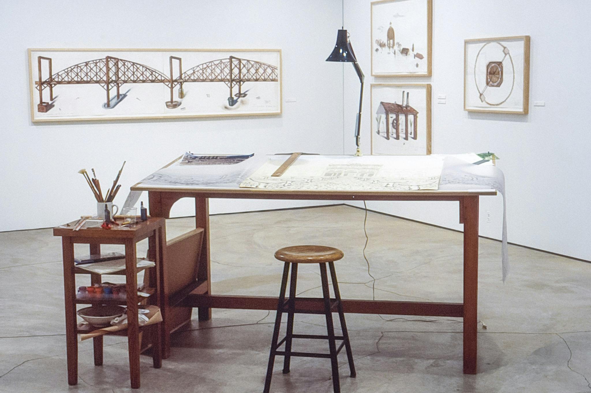 A corner of the gallery resembles the studio space of an architect. Large wooden table with a desk lamp is installed in front of the drawings of buildings and bridges on the walls. 