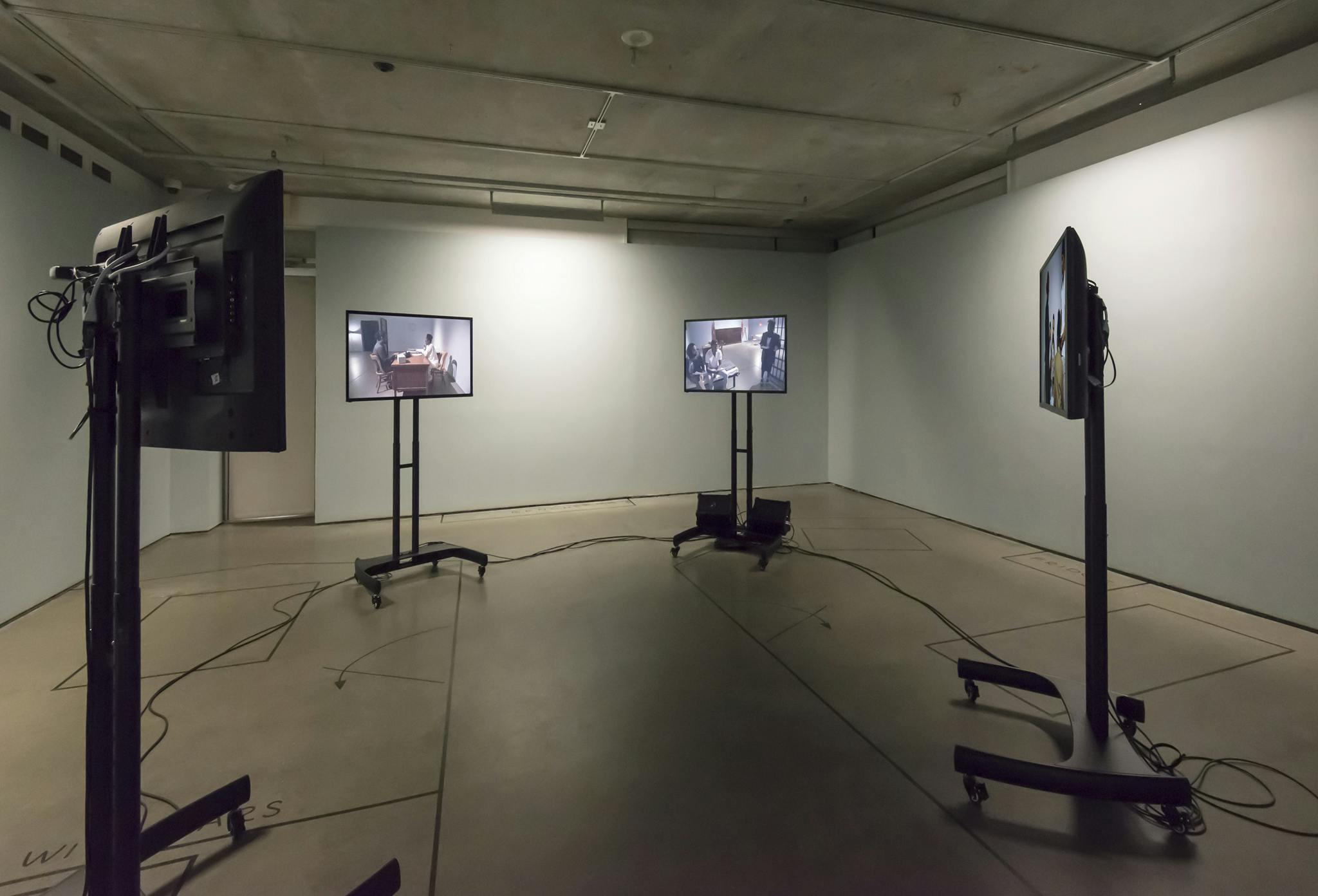 Four monitors are installed in a circle in a gallery. They are all on stands, with the screens facing inwards. Cords and black tape lines lay on the concrete floor. 