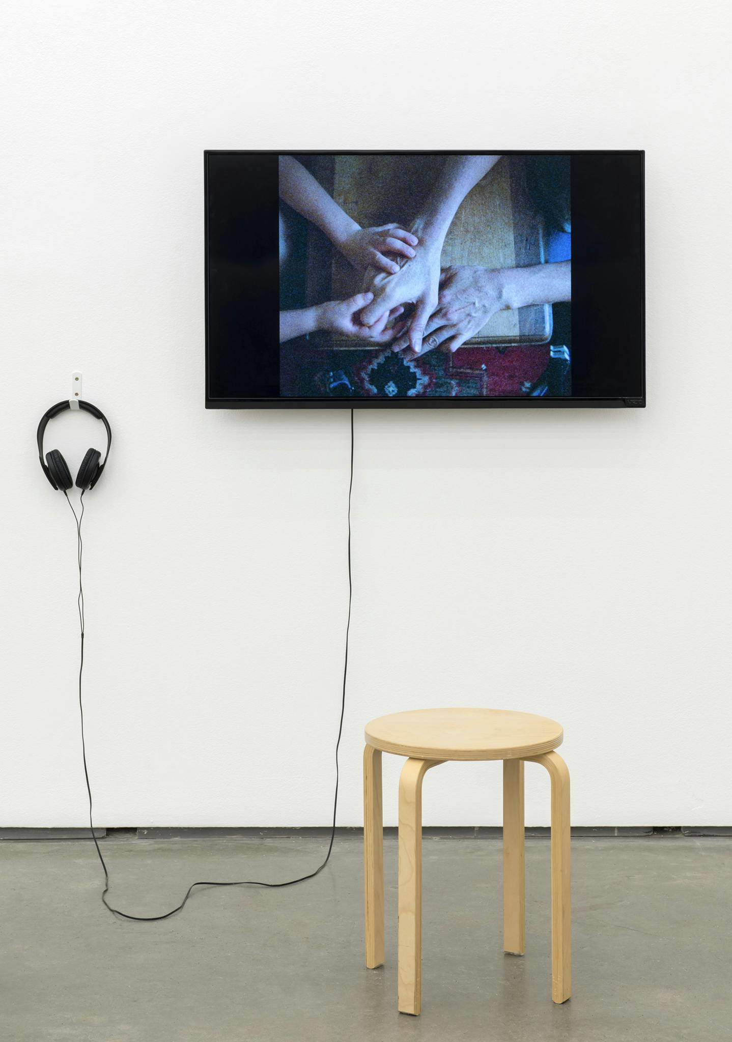 A TV monitor hangs on the wall with a pair of headphones hanging beside it to the left. There is a wooden stool in front of the monitor. The video  on the screen shows four hands holding each other.  