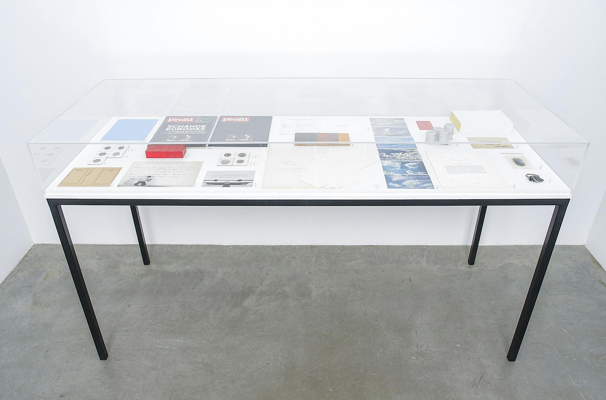 An installation image of artworks by Micah Lexier. A variety of objects, such as photographs of the blue sky, postcards, and magazines are placed in a display case. 