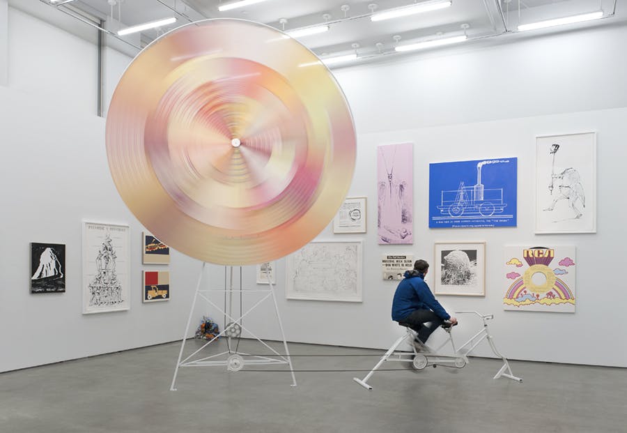 Multiple two-dimensional works of different sizes are mounted on gallery walls behind a large-scale sculpture resembling a wind wheel. The wheel is attached to a bicycle and moves as a person pedals.