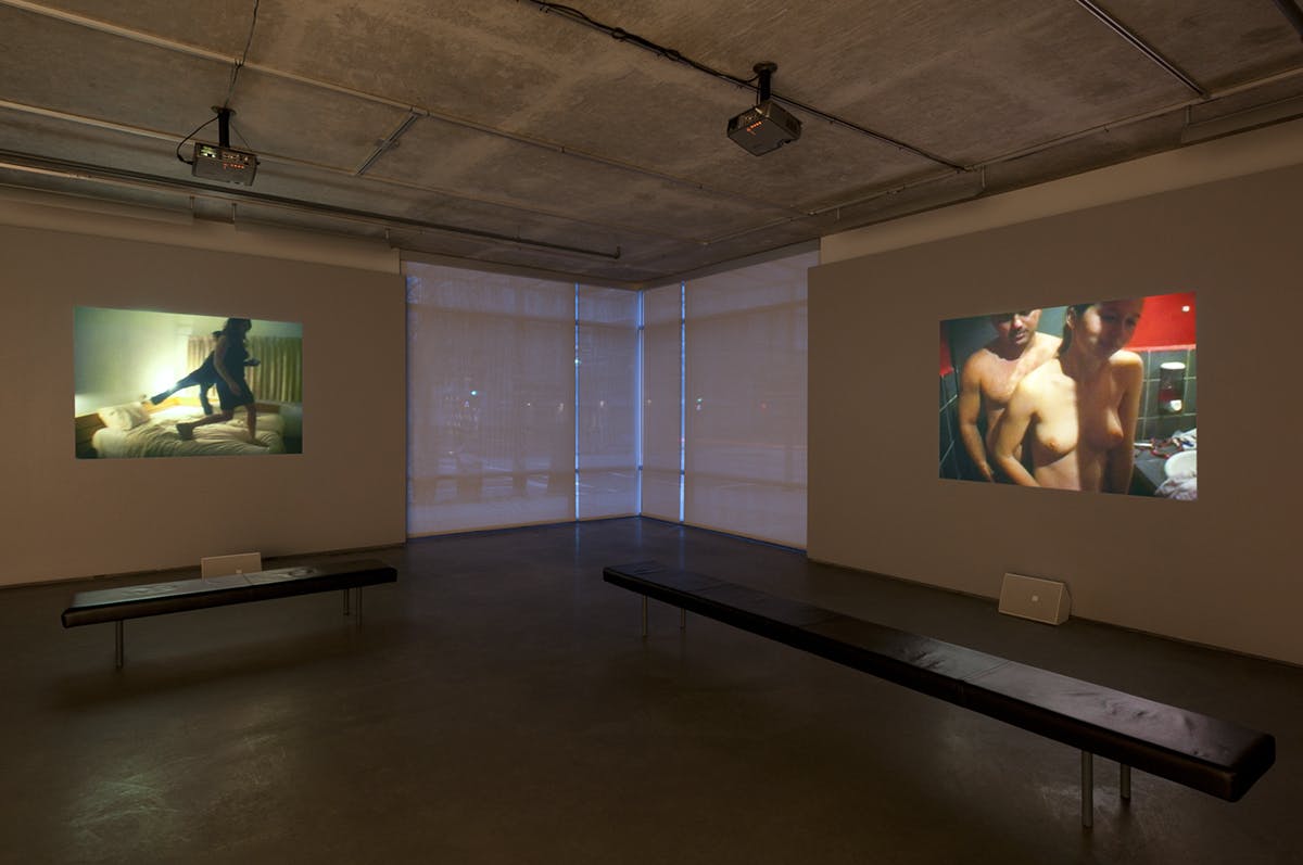 In a gallery, two video works are projected on the walls. The video on the left shows two people in black outfits jumping on a white bed. Another one shows two naked people standing in a washroom.