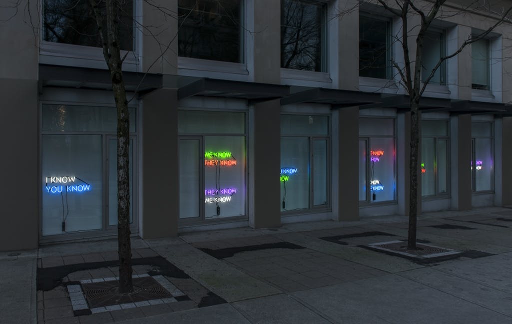 Image of Tim Etchells’ neon text works installed in CAG’s facade windows. Phrases in neon lights of various colours read: “I KNOW YOU KNOW,”“WE KNOW THEY KNOW,””THEY KNOW WE KNOW.”