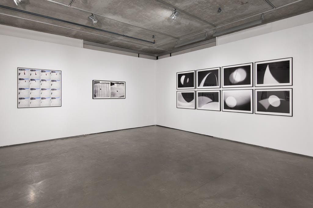 A set of eight black and white abstract photographs are mounted on a gallery wall. Two framed, photographic works are mounted on the adjacent, left-hand wall.