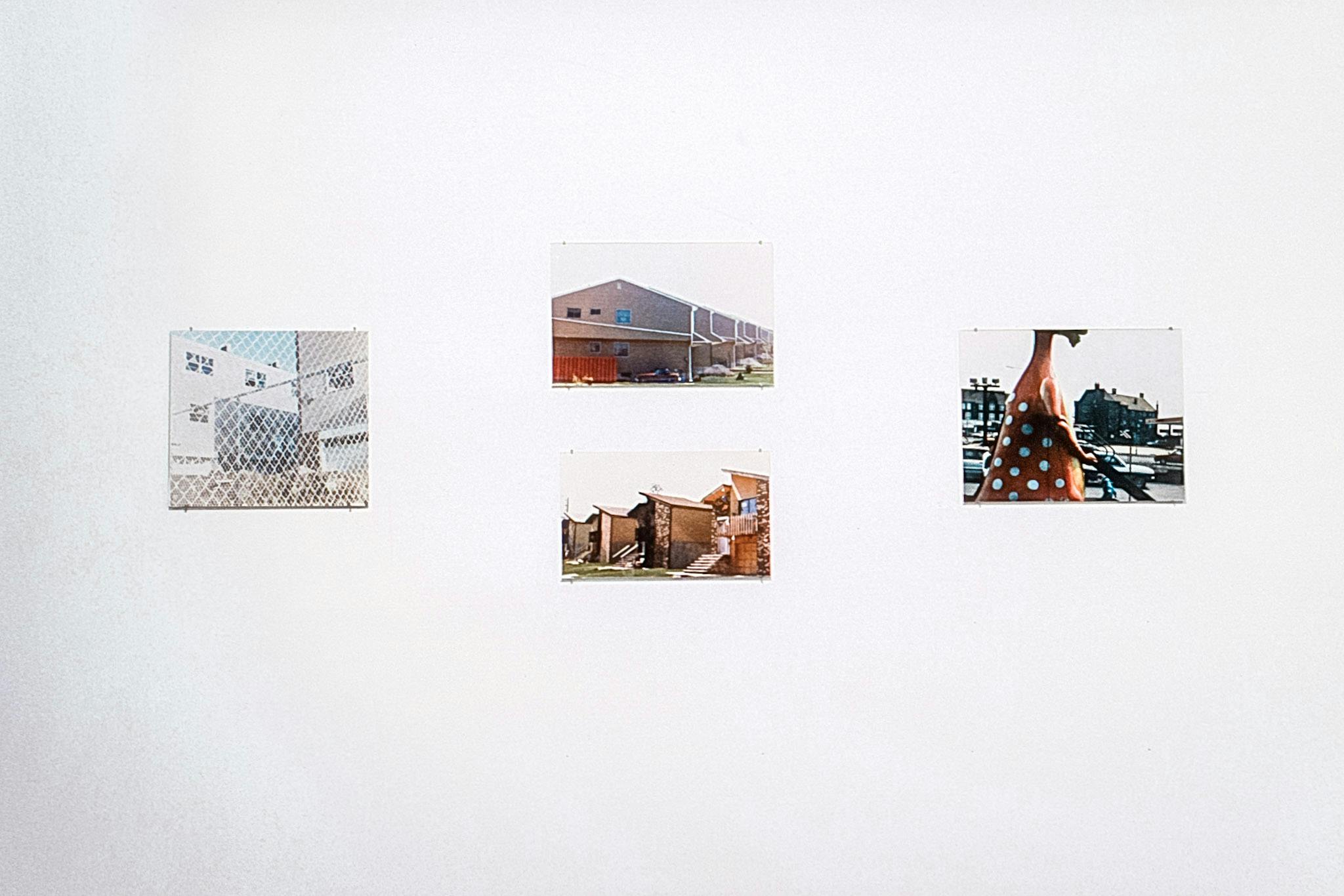 A closeup of 4 unframed photos on a white wall. One has an apartment complex with a fence, one shows townhouses, one has similar houses in a row, and one shows a playground in front of apartments.