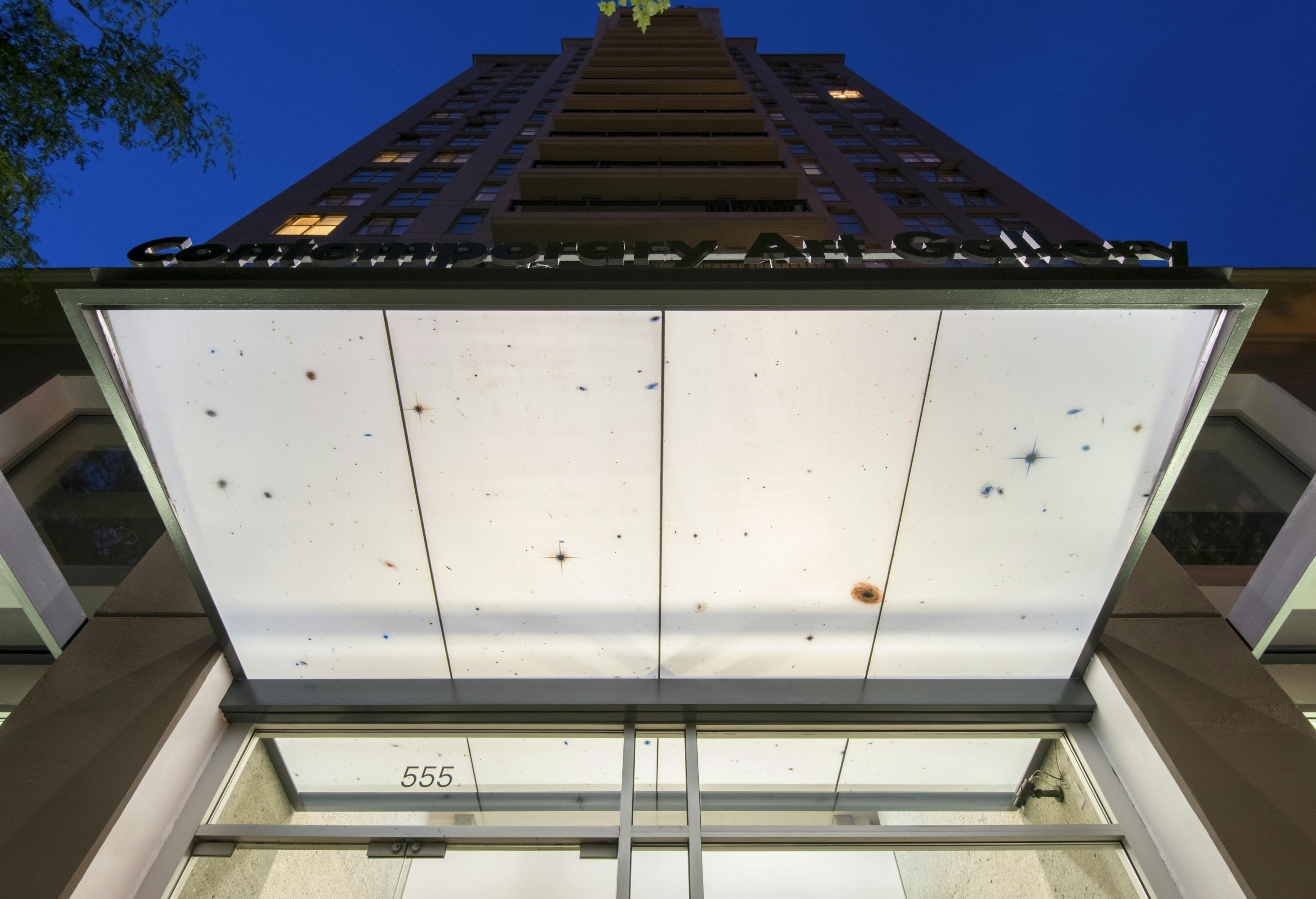 In the glass overhang above CAG’s entrance, four rectangular white panels are installed. Small stars and galaxies in blue, orange, and grey dot the panels. 
