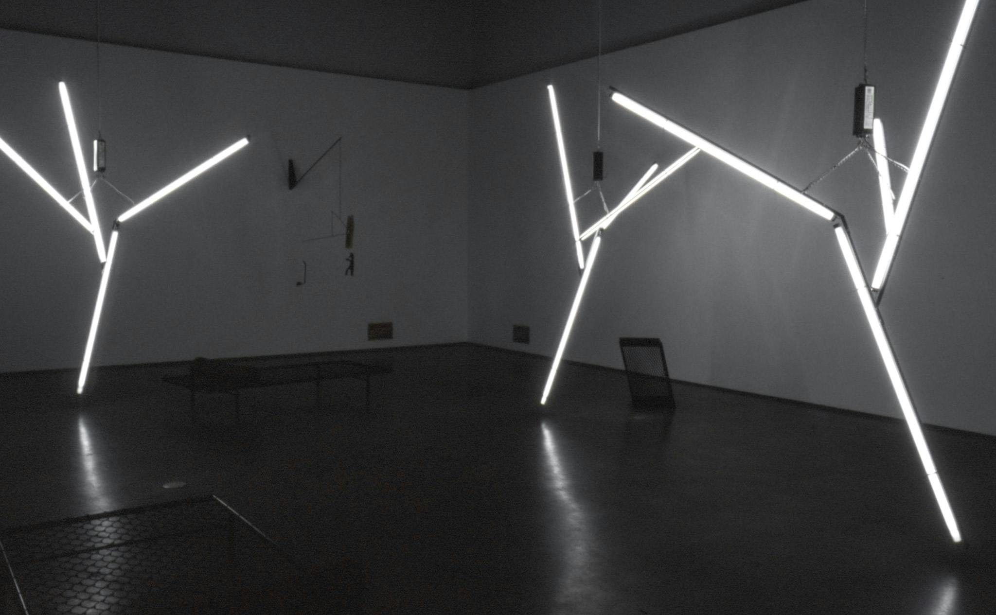 In a darkened gallery space, three large sized sculptures are hanging from the ceiling. They are made of white tube light bulbs that are assembled to look like tree branches. 