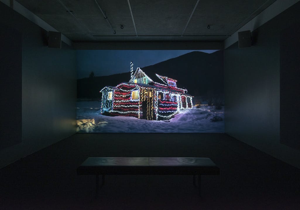 Video projection on a dark gallery wall of a house in a snowy landscape at night. The house is decorated with strings of colourful lights across the exterior walls and roof. Lights are on inside.