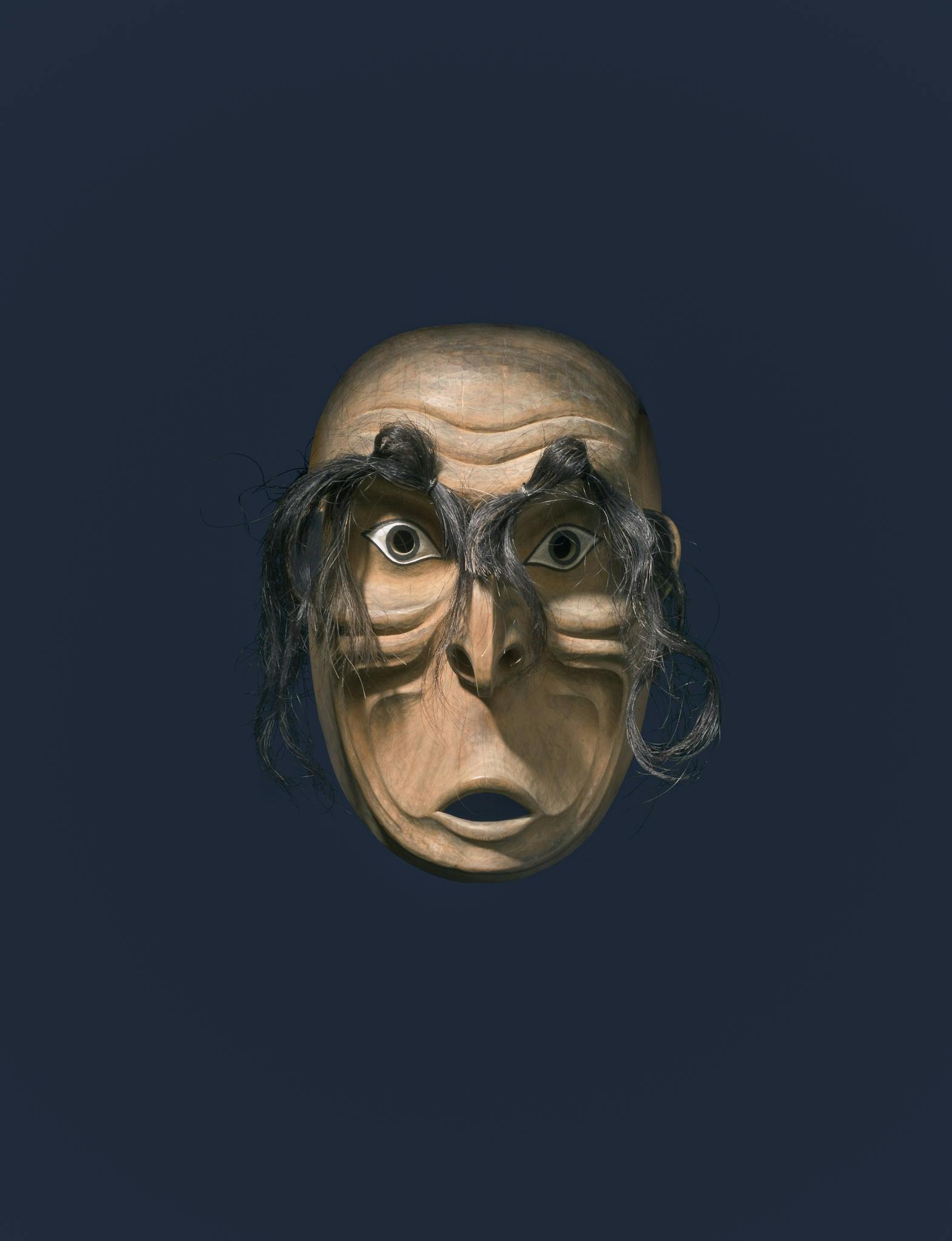 Installation image of Beau Dick’s carved wood mask in front of a black background. The mask has a pair of very long black eyebrows. The mask has a pair of wide-open eyes, a sharp nose, and a mouth opened in a shape of making a “u” sound.