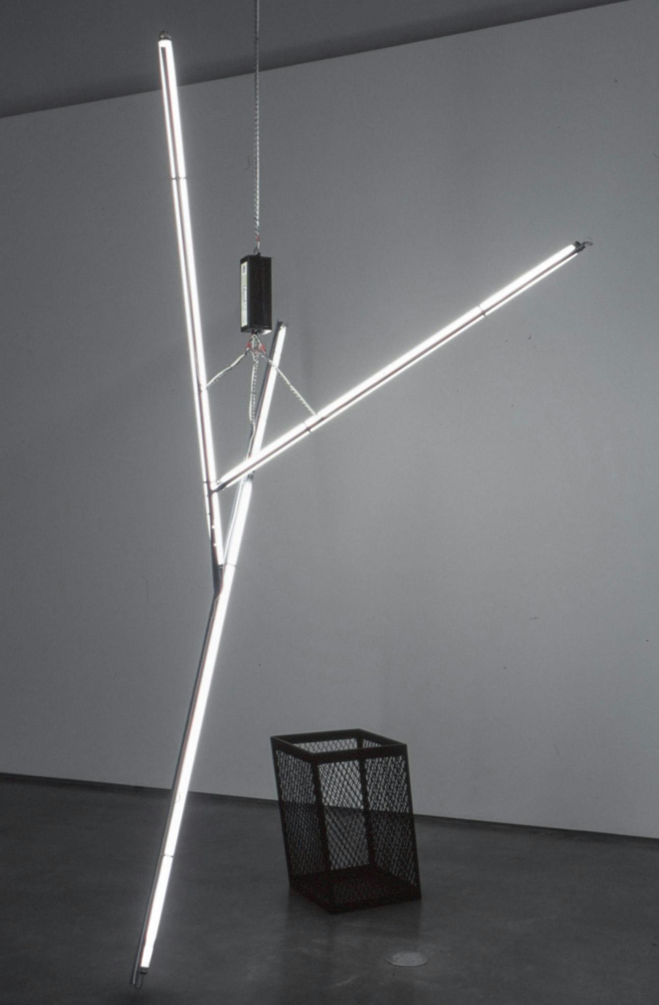 In a darkened gallery space, a large sculpture made of white tube light bulbs is hanging from the gallery ceiling. A black trash can, or an umbrella bucket, is placed behind this large sculpture. 