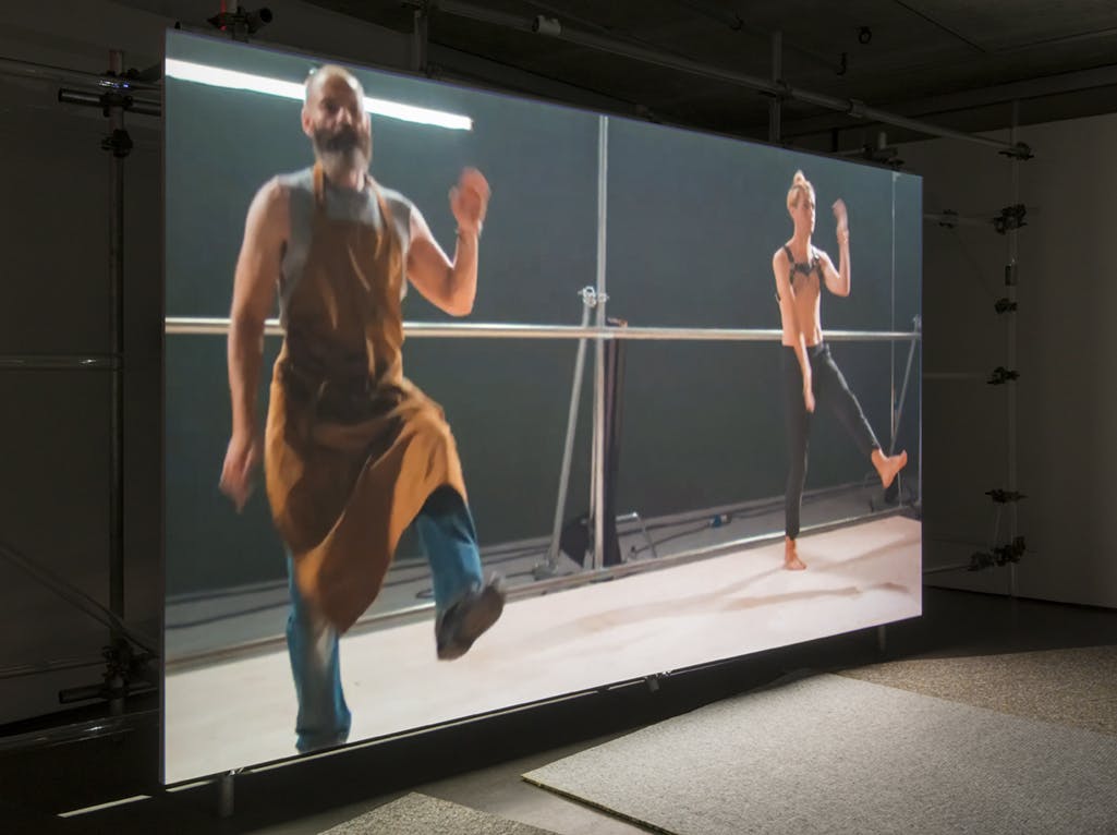 A video work by Patrick Staff is projected onto a large screen in a darkened gallery. Two figures are dancing the same choreography. 