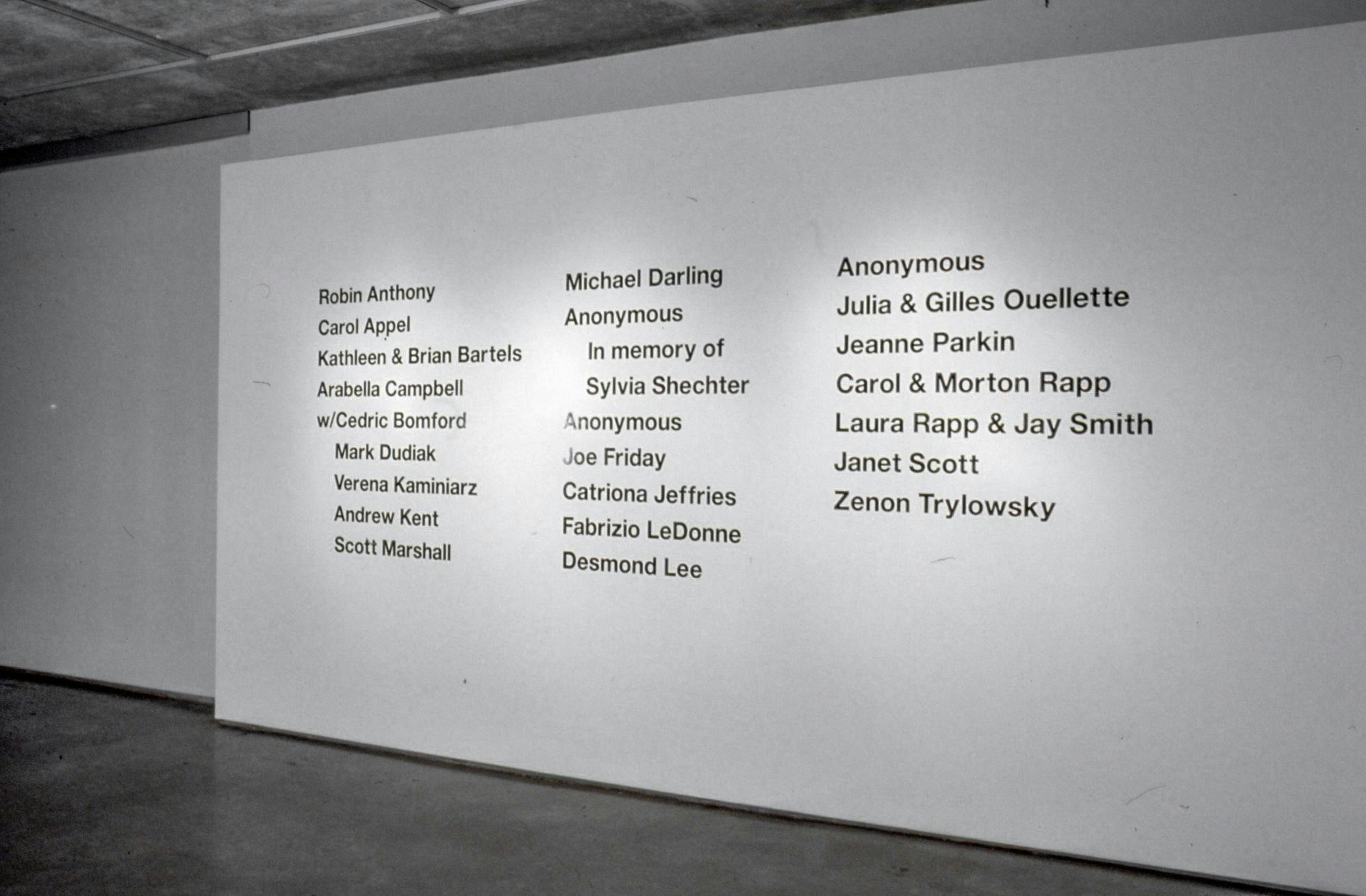 This is an installation shot of a gallery space. On a white wall, the names of people are printed in a large black font. There are two listed as “Anonymous” in this list of names. 