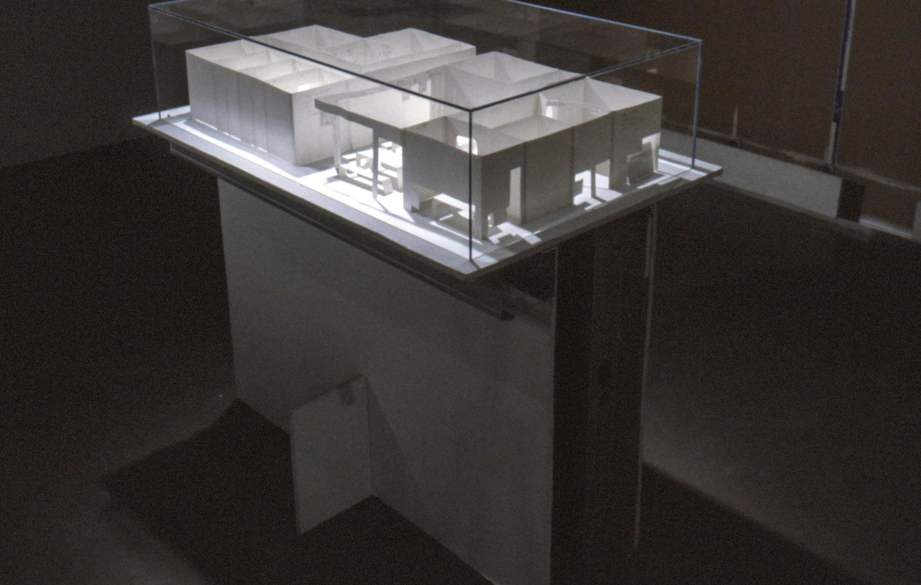 In a darkened gallery space, a white mockup of an architecture complex is displayed. This sculpture sits on a part of a white wall which has been removed from one of the galley walls.  