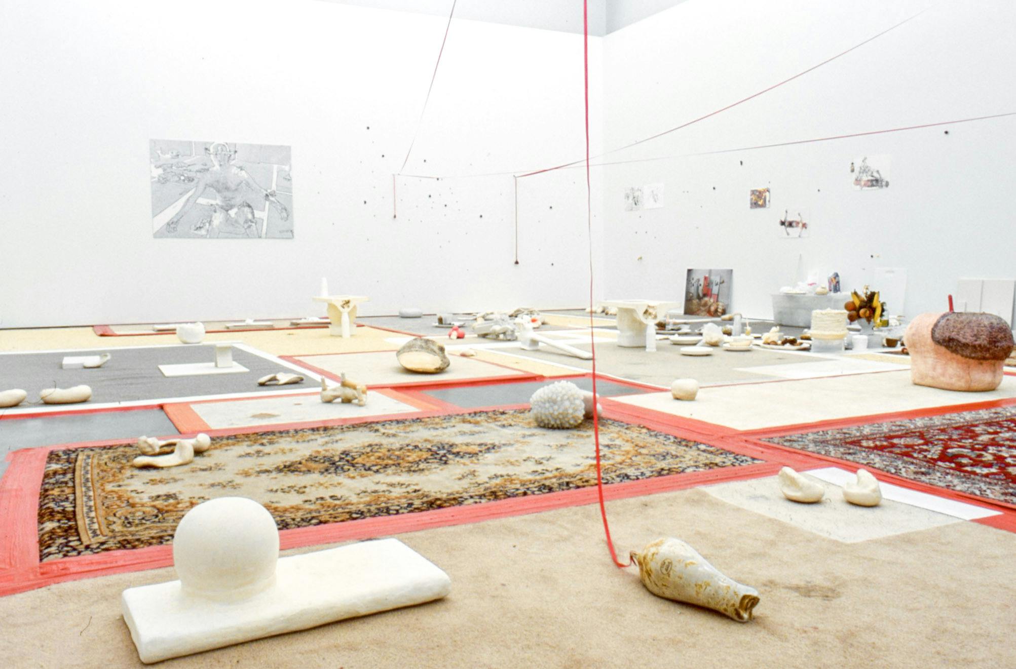 Installation image of artworks in a gallery. A variety of found objects, including shellfish and glass bottles are placed on the floor. The floor is partially covered with the patchwork of various carpets.