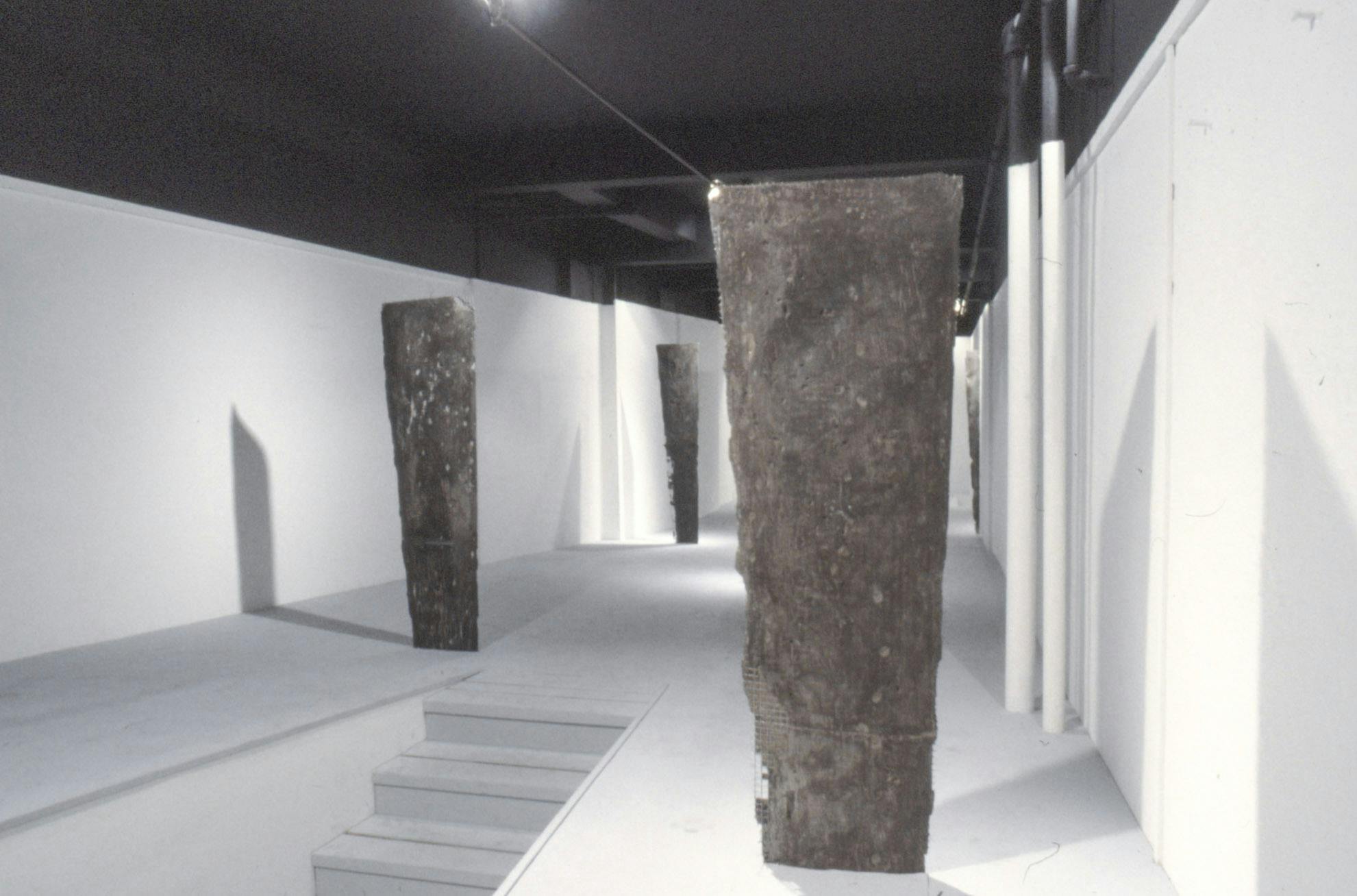 Several vertical structures made of cast metal-cement and wire stand on a white surface in a gallery, close to the ceiling. A set of steps leading to the works is partially visible.
