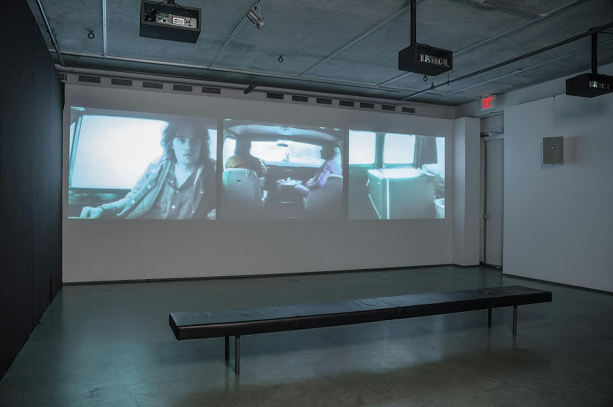 An installation image of three-channel video work projected on a gallery wall. The videos show people and objects in a car from different angles. The middle video shows an image of two people in front seats.