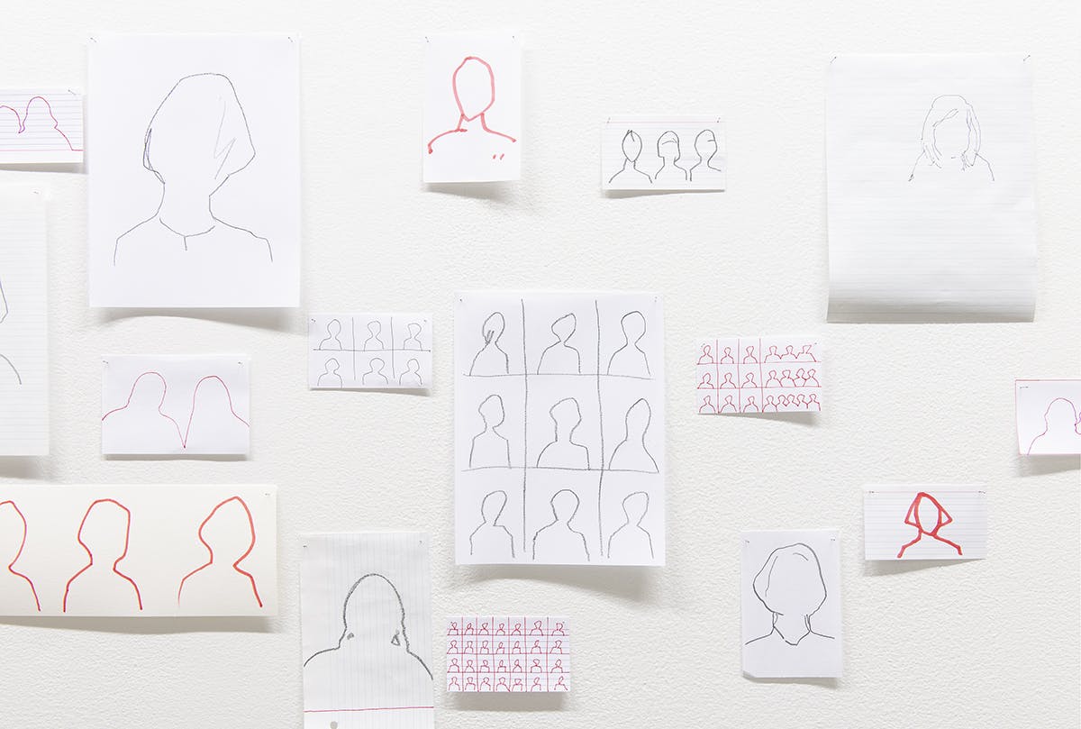 This is a close-up view of Faye HeavyShield’s drawings installed on a gallery wall. They were drawn with several different mediums, including markers and pens. They depict the shapes of human heads.