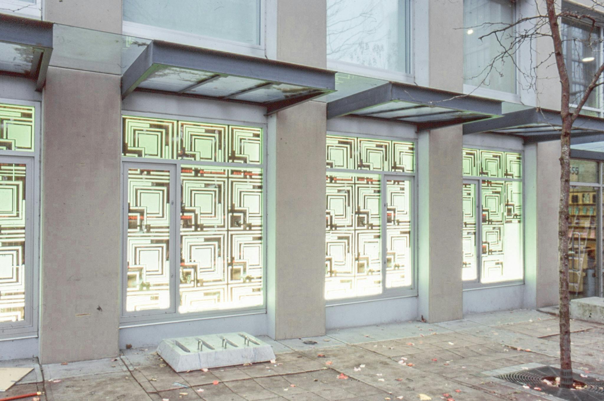 An installation image of artworks in CAG’s window spaces facing a sidewalk. The wall behind glass windows is covered with identically designed posters. The poster’s design is made of a combination of geometric shapes. 