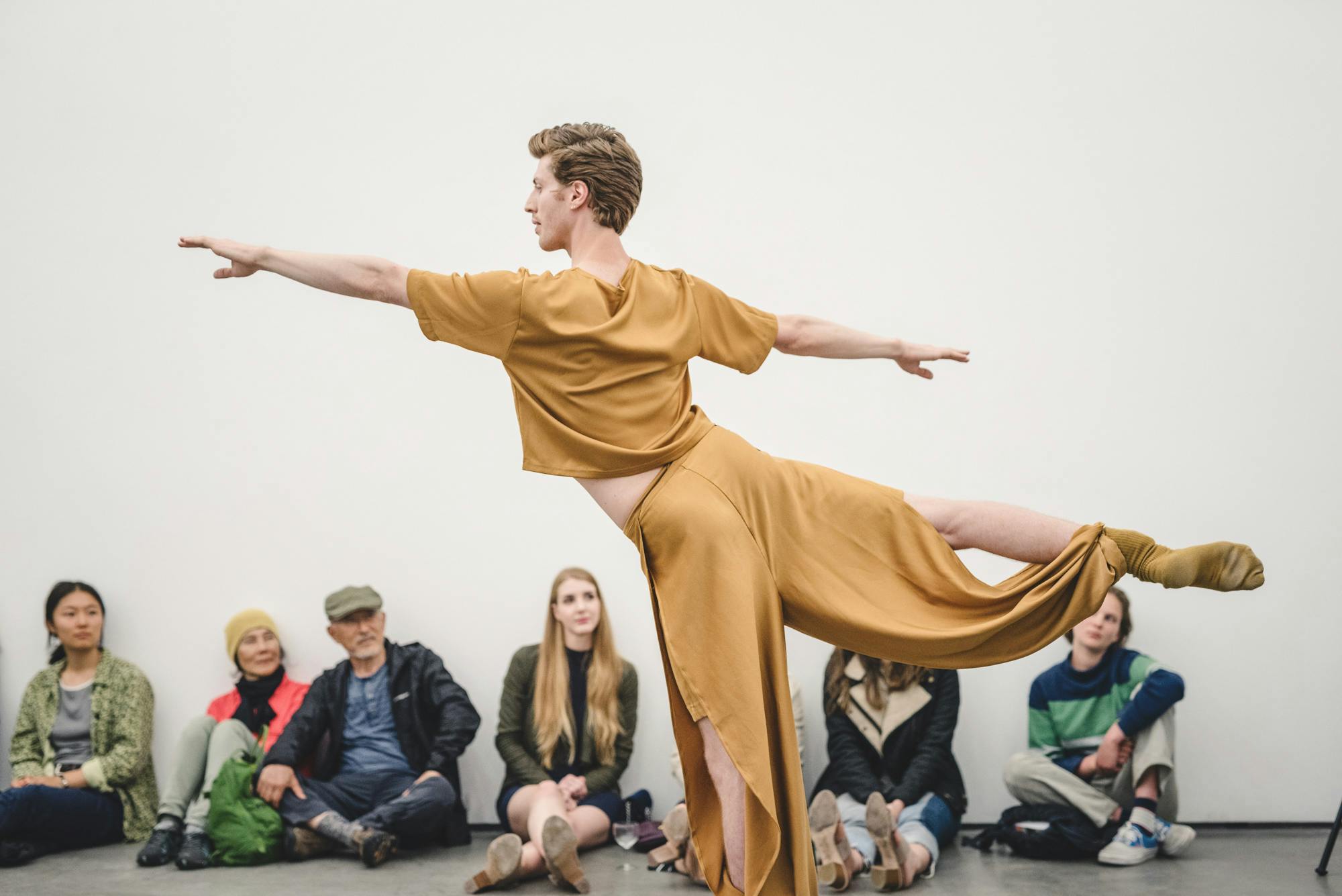 Andrew Bartee, wearing a mustard-coloured outfit is performing for an audience seated on the floor against a gallery wall.