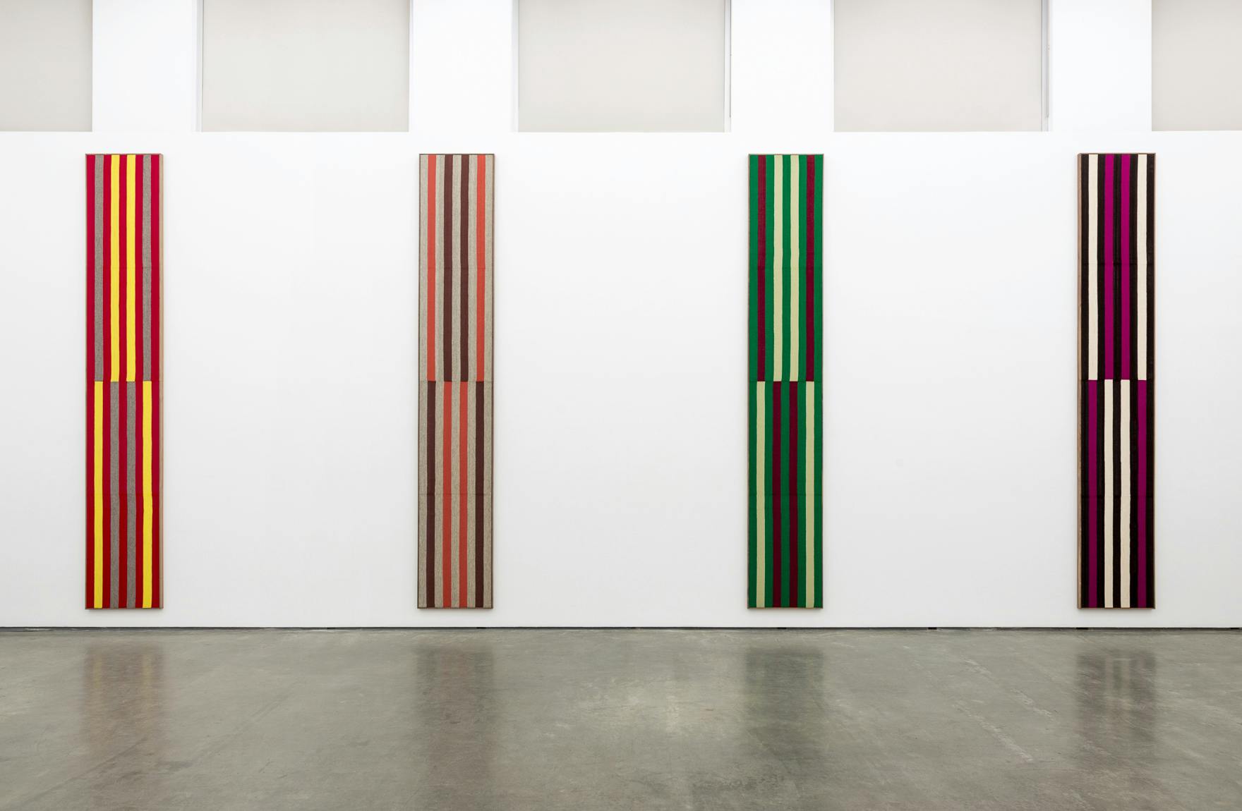 Installation image of Brent Wadden’s exhibition Two Scores. Four striped woven works stretched onto narrow, rectangular panels hang vertically on a gallery wall. 