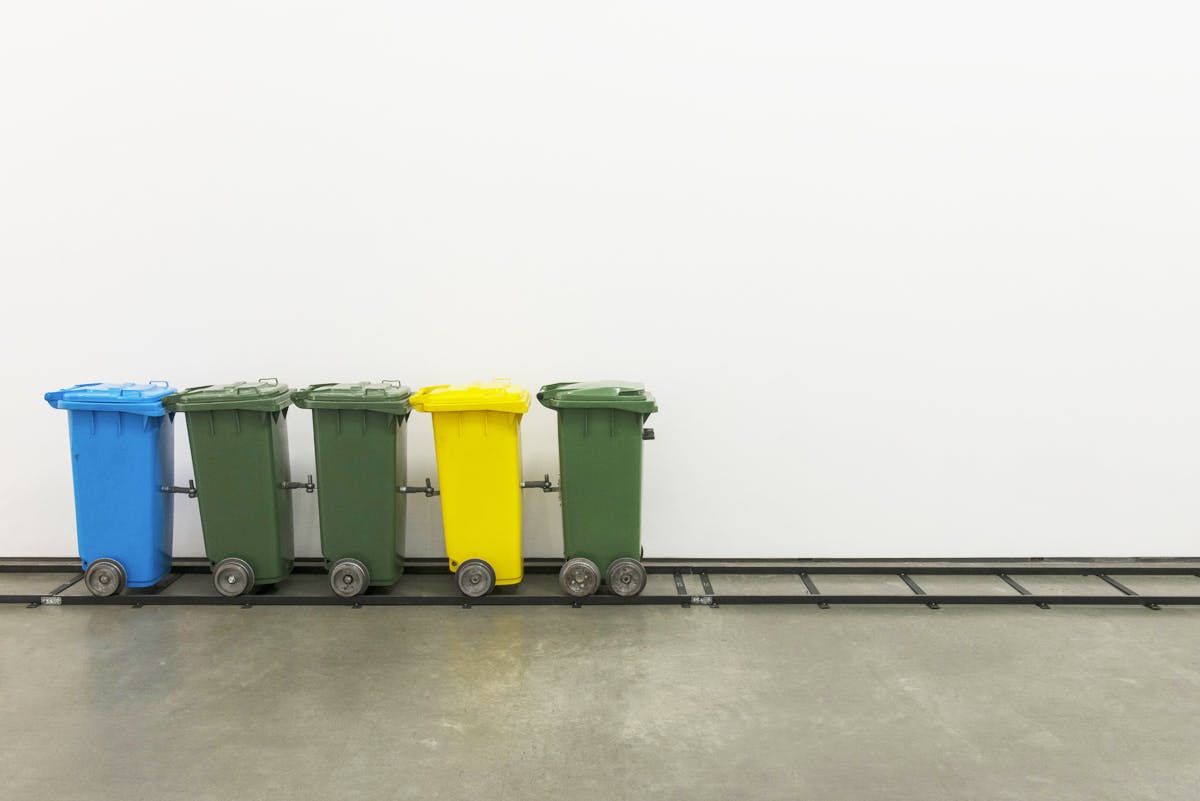 Five garbage bins are connected via metal train couplers. A pair of tires are attached to the bottom of each bin to make them mobile. They align on a black rail built inside a gallery space.